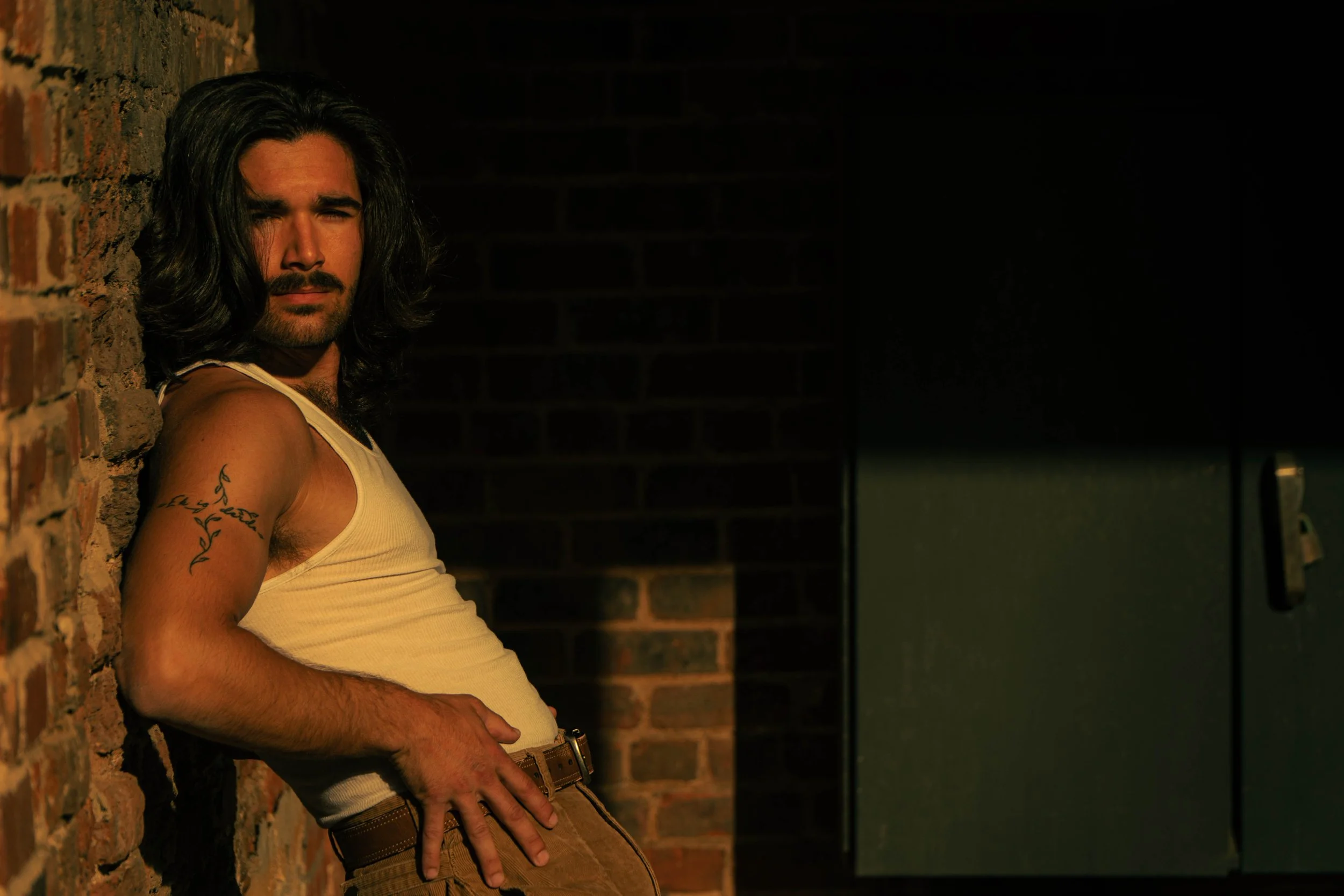 A man with long dark hair and a beard leaning against a brick wall, wearing a white tank top and tan pants, in a dimly lit setting.