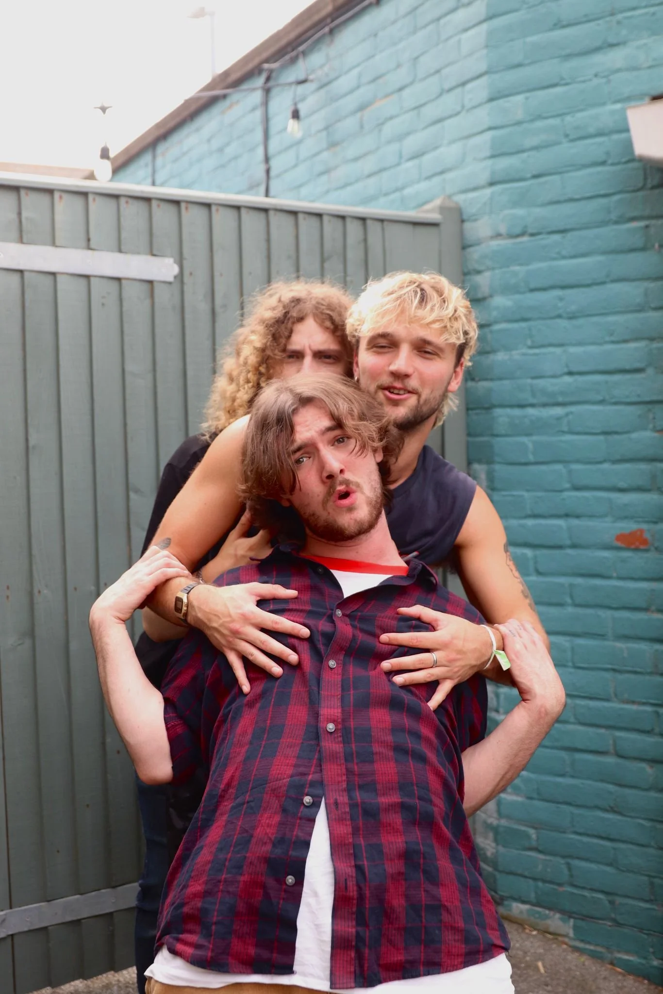 Three young men are close together, posing playfully outdoors in front of a blue brick wall and a gray fence. The man in front has brown hair, a beard, and wears a red and black checkered shirt. The man behind him has blonde hair, a light beard, and is wearing a sleeveless shirt. The third man, with curly hair, is slightly behind and has a surprised expression.