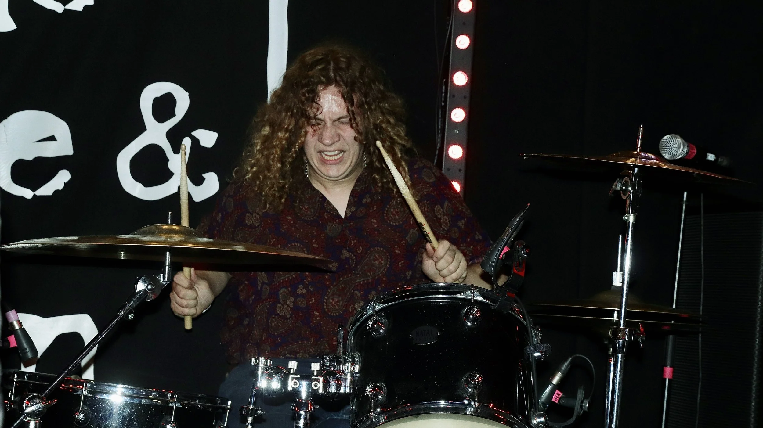 A woman with curly hair playing a drum set on stage, with a signature in the background and stage lights.