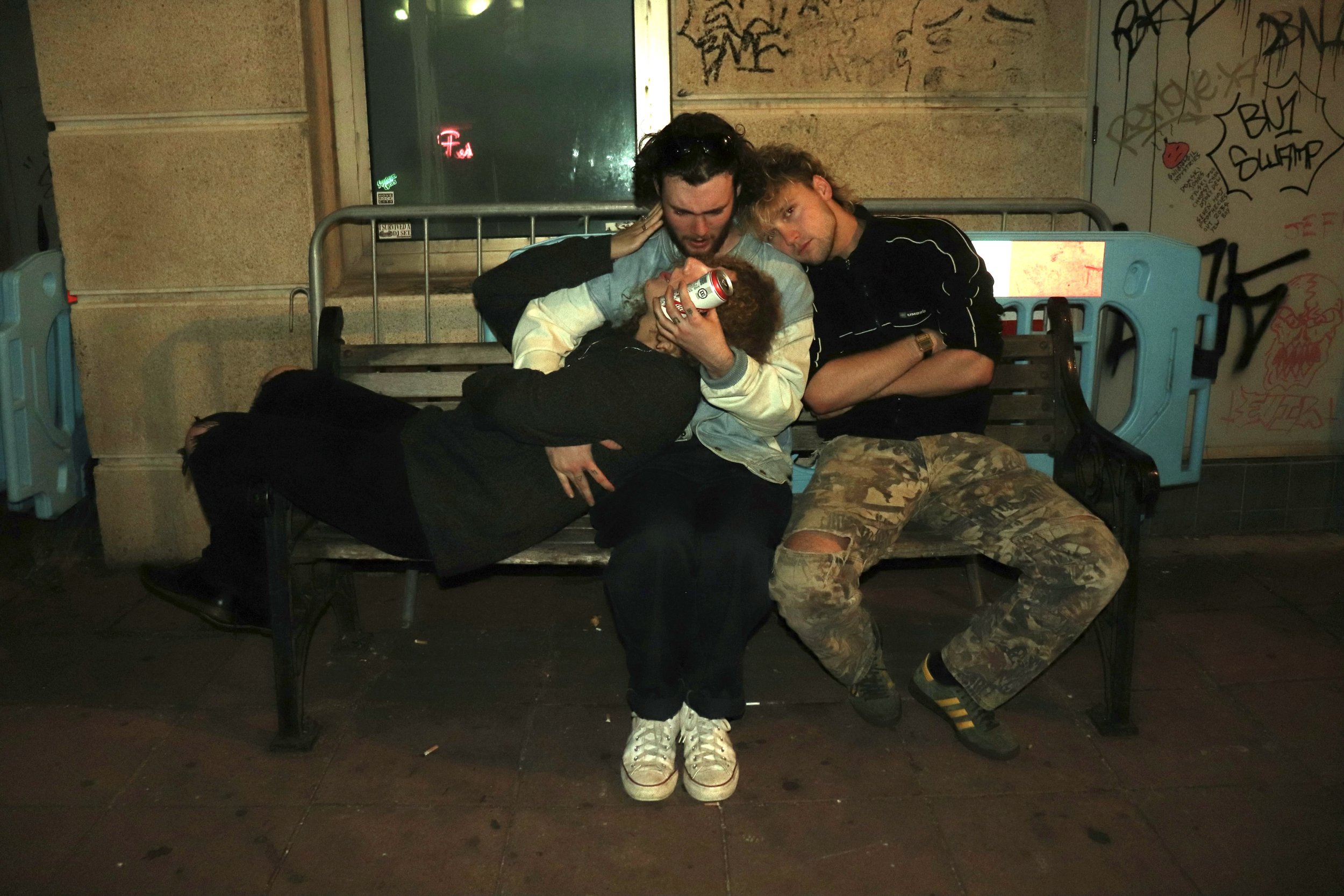 Four young people sitting on a park bench at night, with graffiti on the wall behind them. They are relaxed and socializing, one holding a can of beer, others leaning on each other.