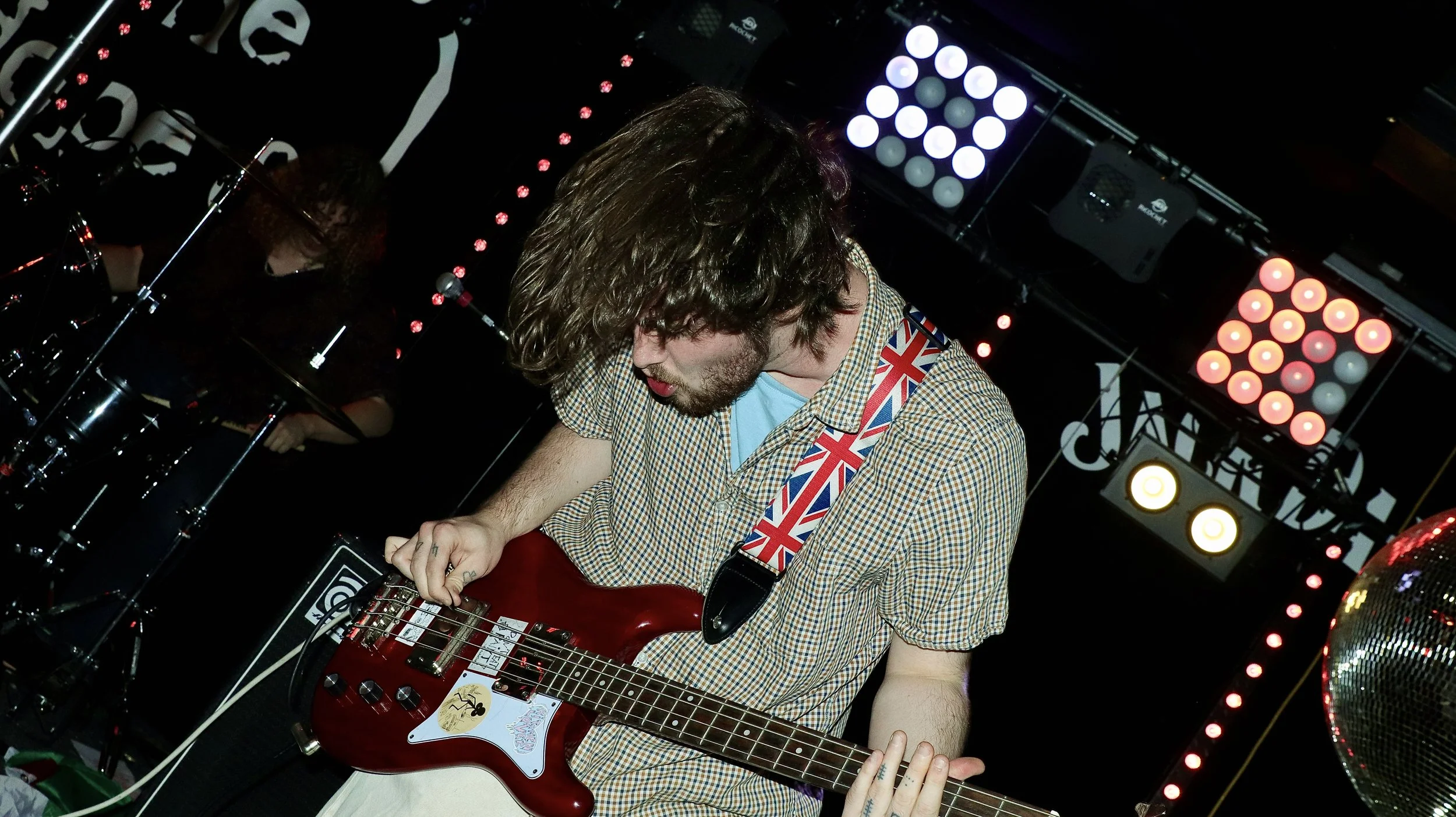 A musician with long curly hair playing a red electric bass guitar on stage, wearing a checkered shirt with a Union Jack guitar strap, with colorful stage lights in the background.