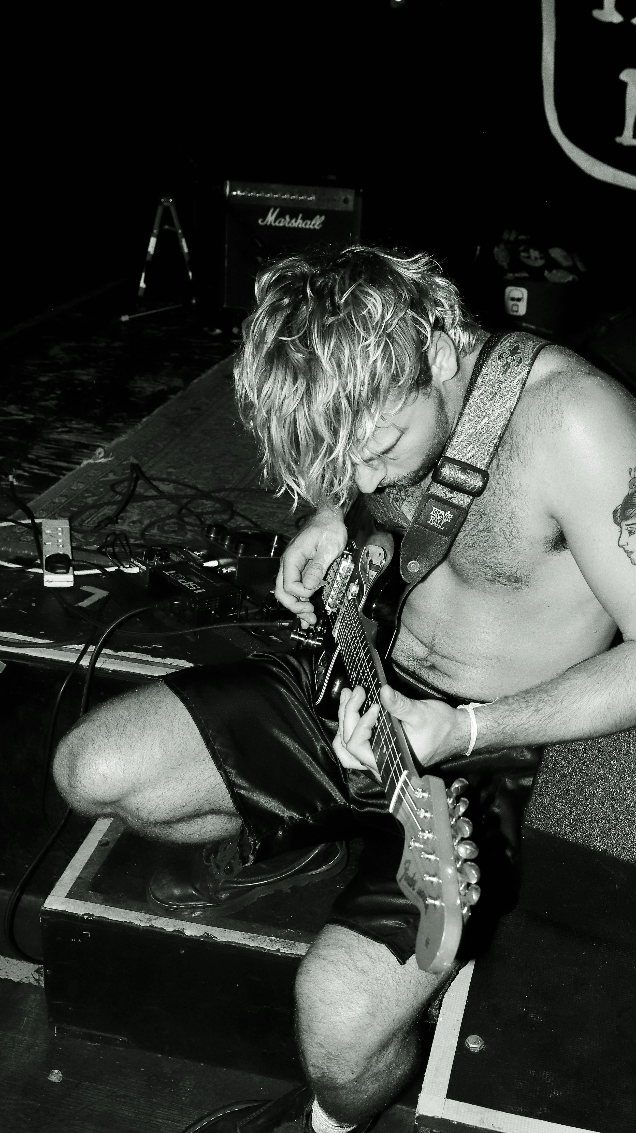 A shirtless man with long curly hair playing an electric guitar on stage, with music equipment and a Marshall amplifier in the background.