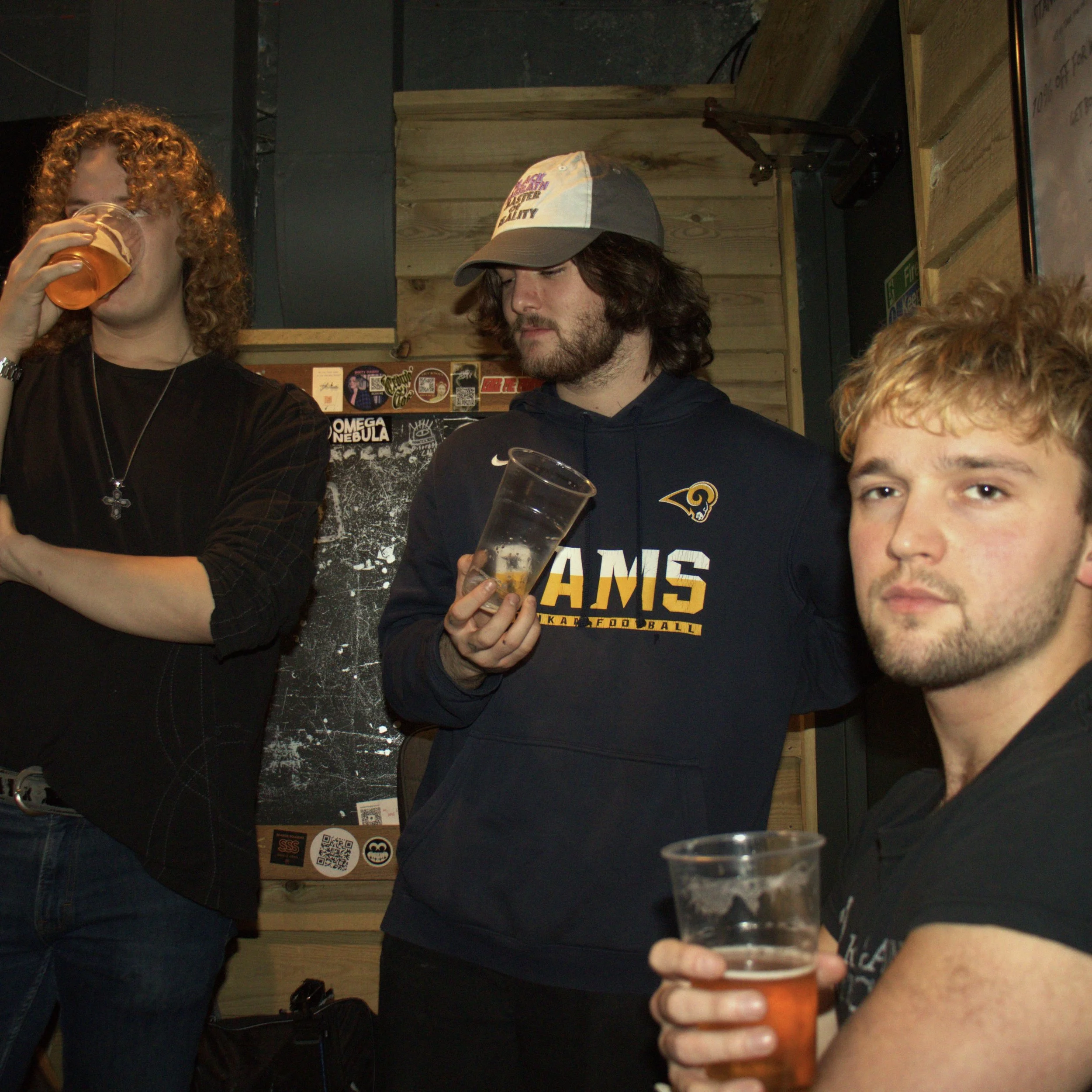 Three young men with light skin are at a bar, holding drinks. The man on the left is drinking from a glass with beer, has curly red hair, and is wearing a black shirt with a necklace. The man in the middle has wavy dark hair, a beard, and is wearing a baseball cap and a hoodie with the logo of the Los Angeles Rams football team, holding a glass of beer. The man on the right has blond hair, a short beard, and is holding a beer, looking at the camera.