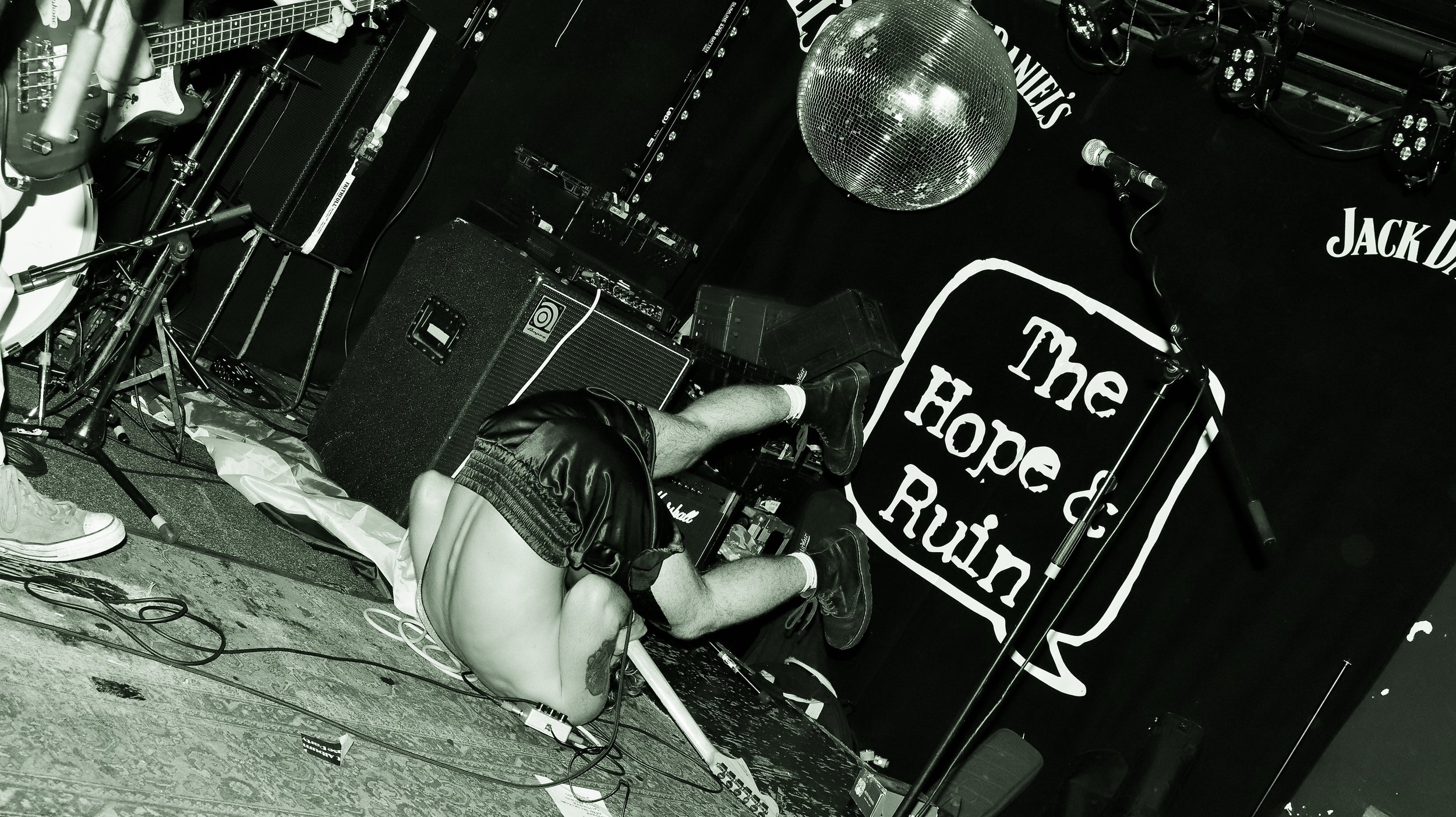 A person lying upside down on stage with their head and arms on the ground, legs raised against a speaker. They are shirtless, wearing shorts and sneakers, with a tattoo visible on their arm. The stage has musical equipment, including a guitar, microphone, amplifiers, and a disco ball. The backdrop features a sign with the phrase 'The Hope & R.....' and the logo 'Jack J...'.