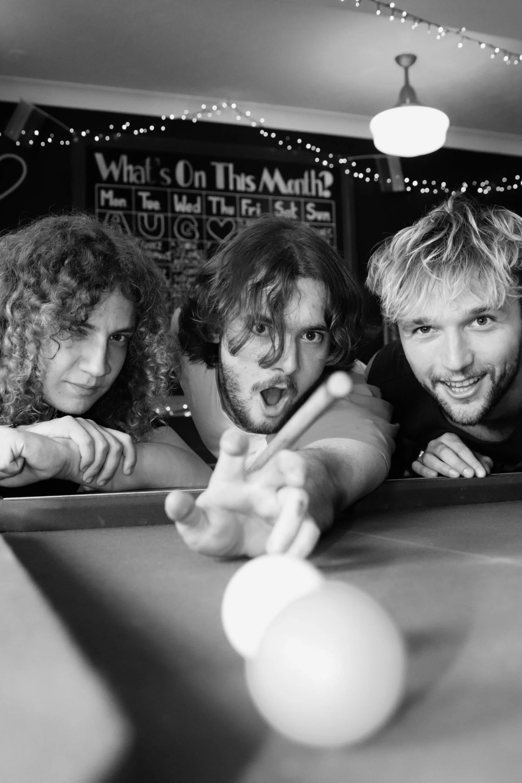 Three friends playing pool, with one making a shot toward the camera, in a bar with string lights and a calendar on the wall.