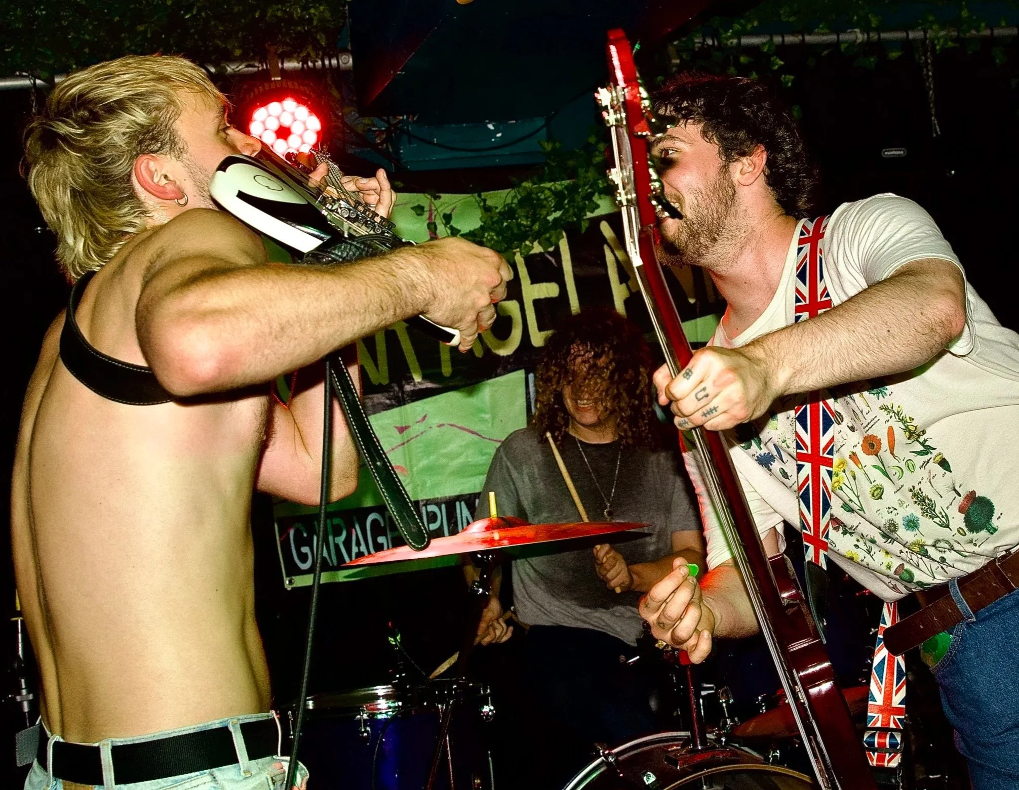 Two male musicians playing guitars and singing into each other's faces on stage, with a male drummer in the background, at a lively music venue. A Brighton punk band playing live at Daltons.