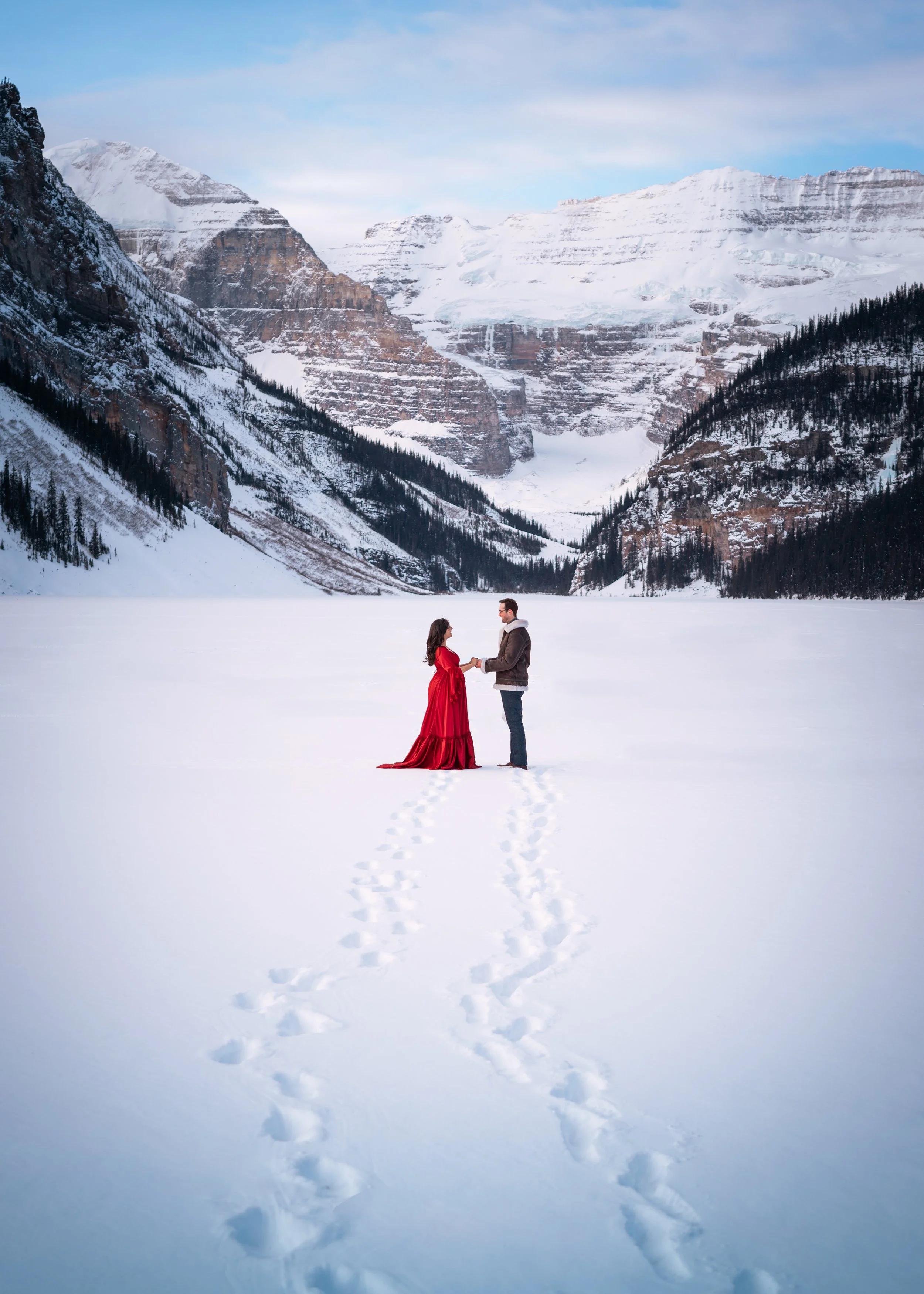 A couple stands in the snow holding hands, with mountains and trees in the background.