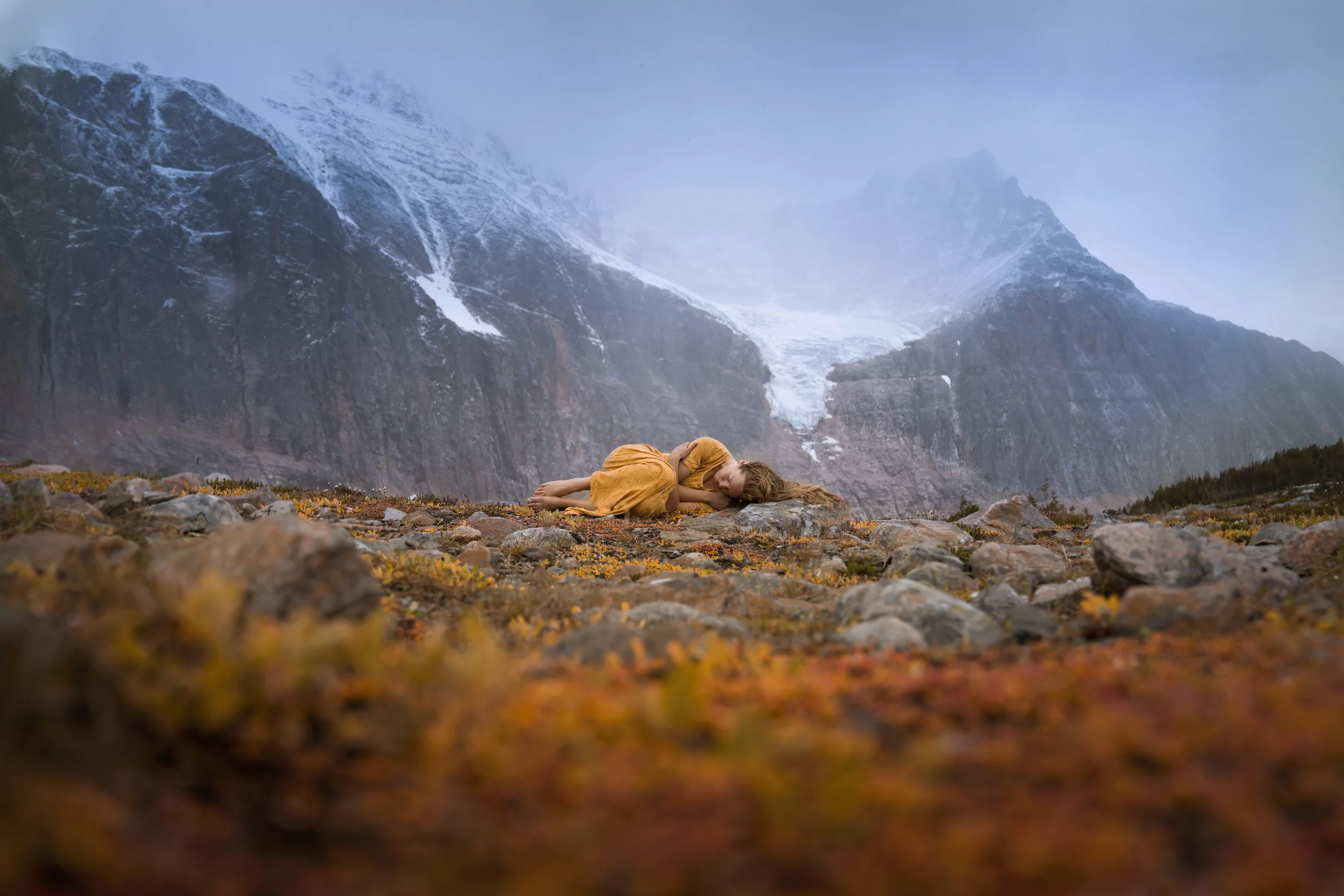 A woman in a yellow dress lying curled up on the ground among rocks and low plants in a mountainous landscape with snow-capped peaks in the background.