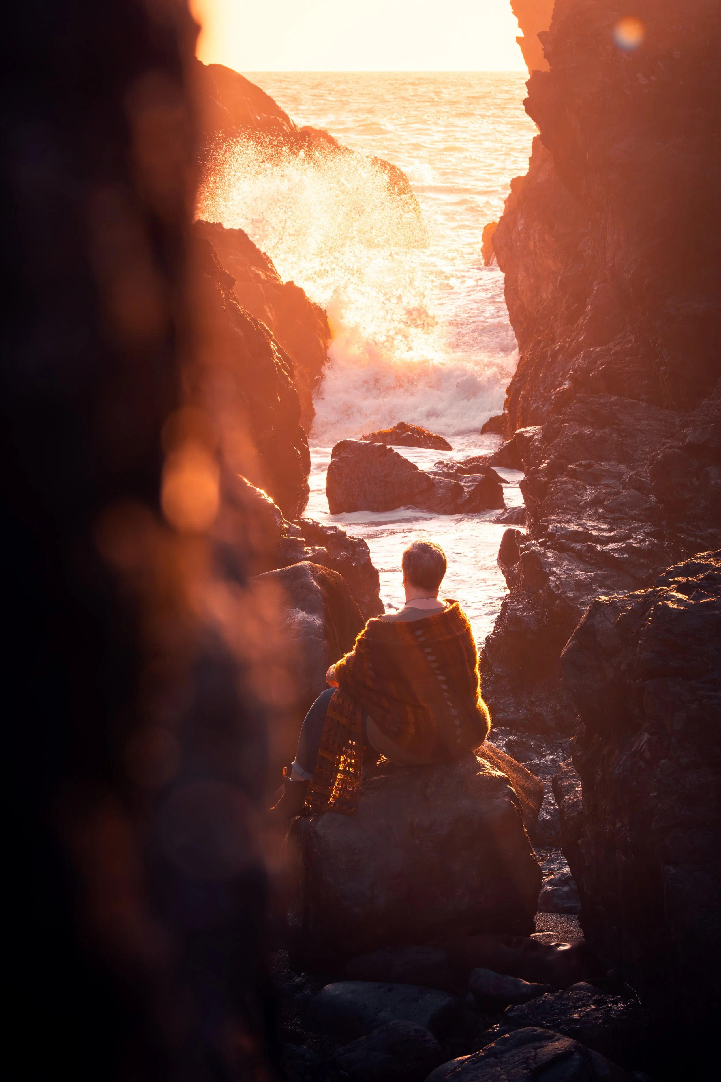 A person sitting on rocks between cliffs, looking at the ocean during sunset, with waves crashing and the sun casting a warm glow.