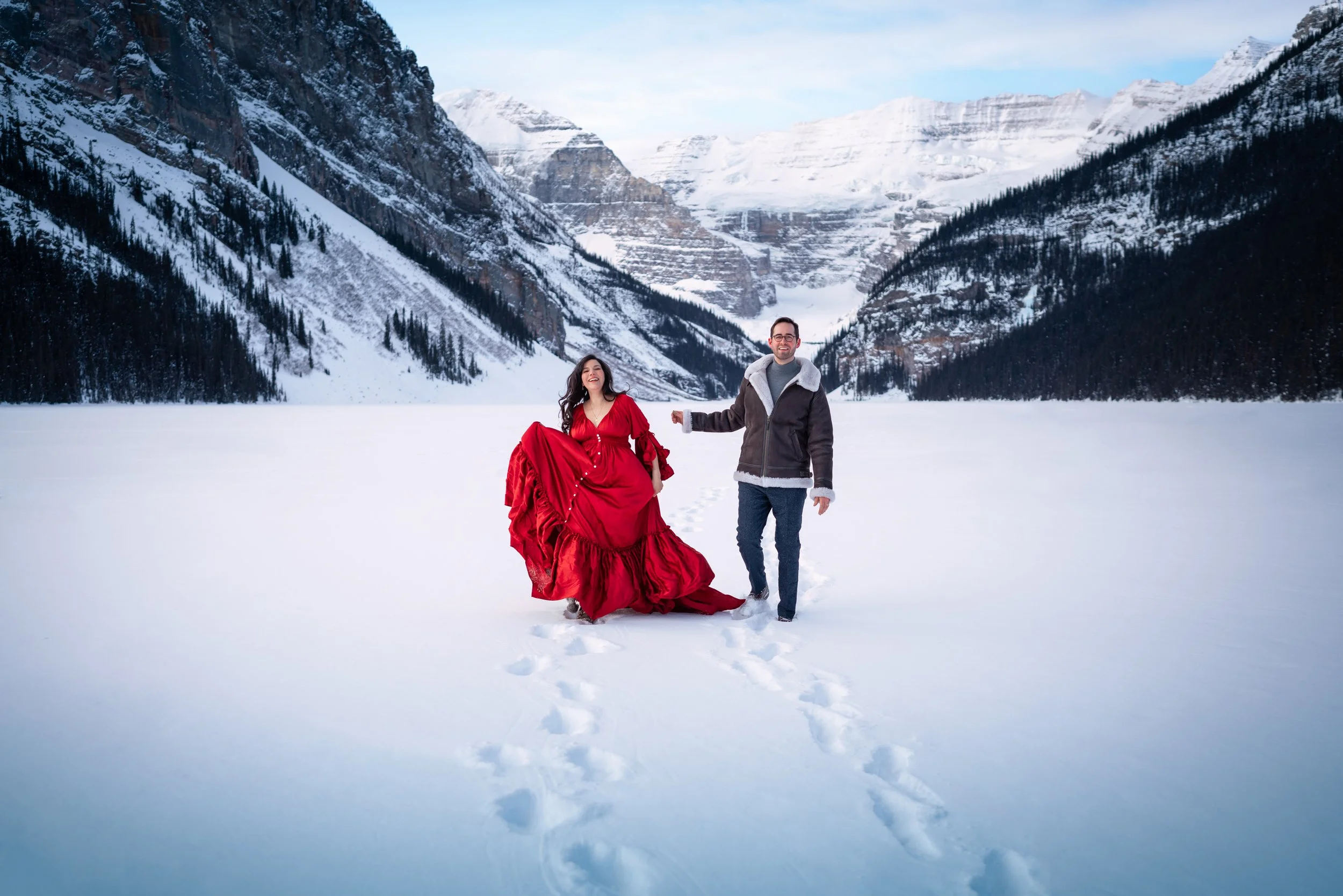 A man and woman walking in a snowy mountain landscape; the woman is wearing a long red dress and the man a brown jacket, both smiling and holding hands, with snow-covered mountains in the background.