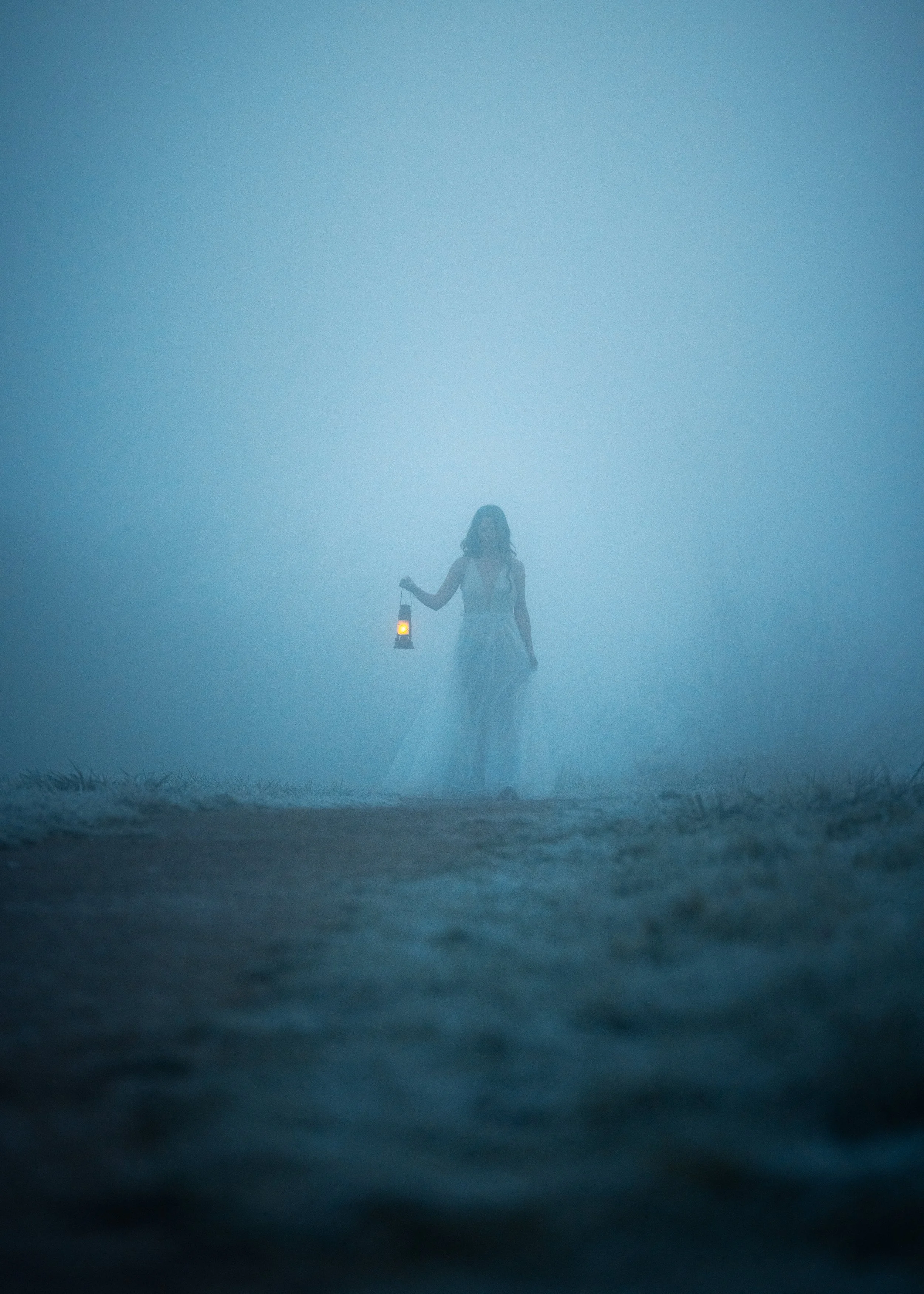 A woman in a white dress walking through foggy terrain, holding a lantern with a small glowing light inside.