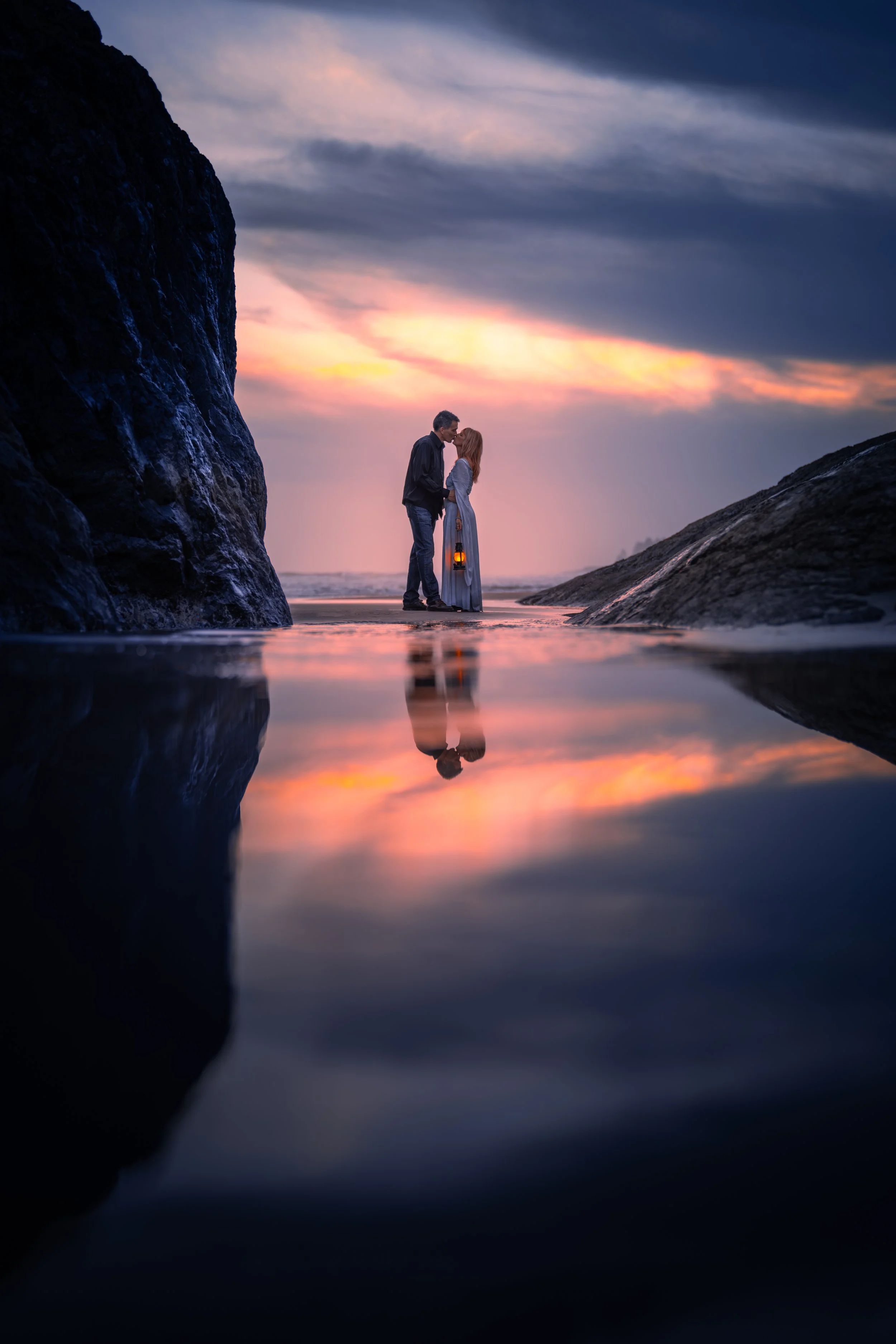 A romantic couple standing on a beach at sunset, holding a lantern, with their reflection visible in the water, framed by large rocks.