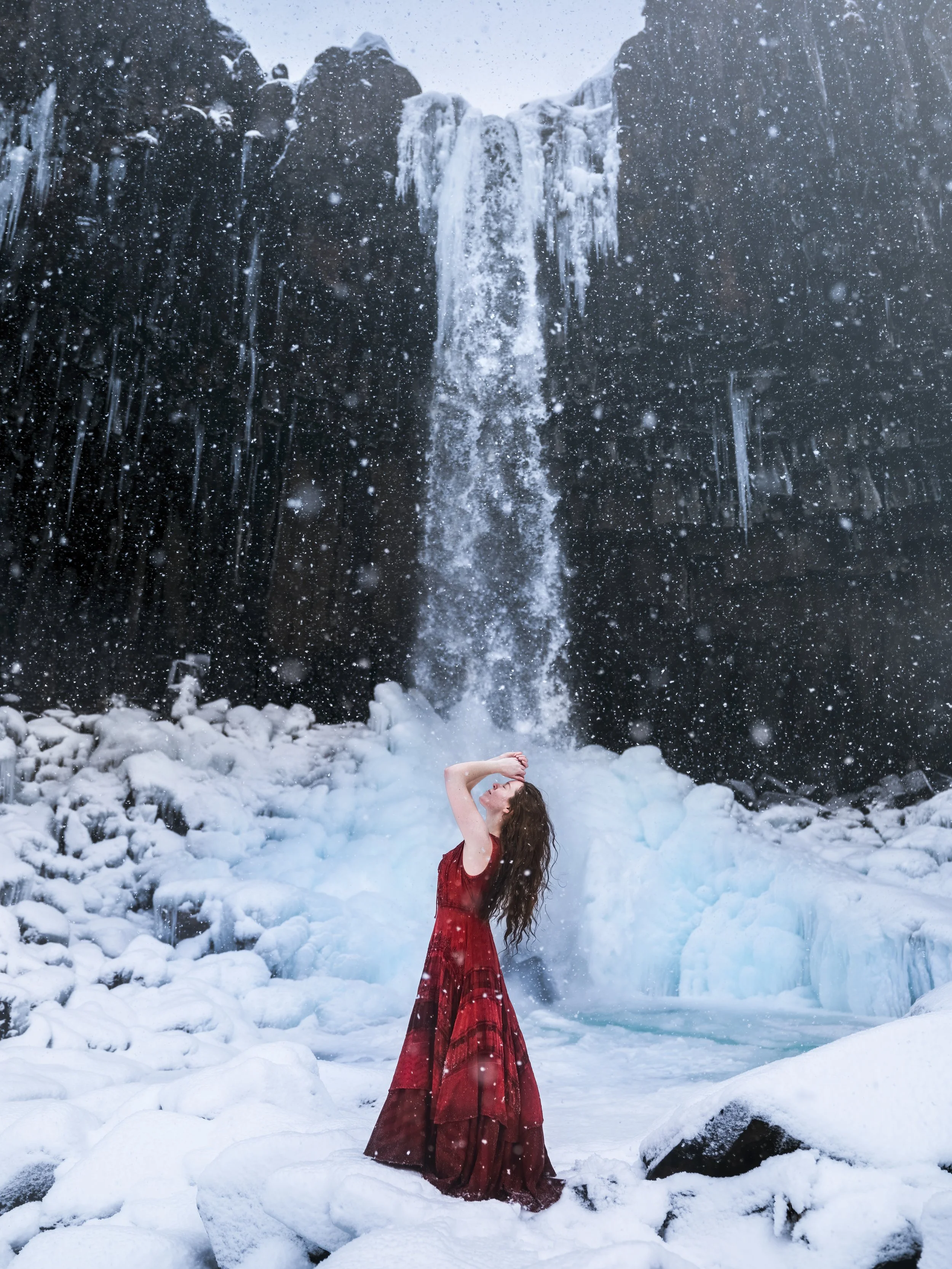 A woman in a flowing red dress stands beneath a waterfall surrounded by snow and ice, with snow falling around her.