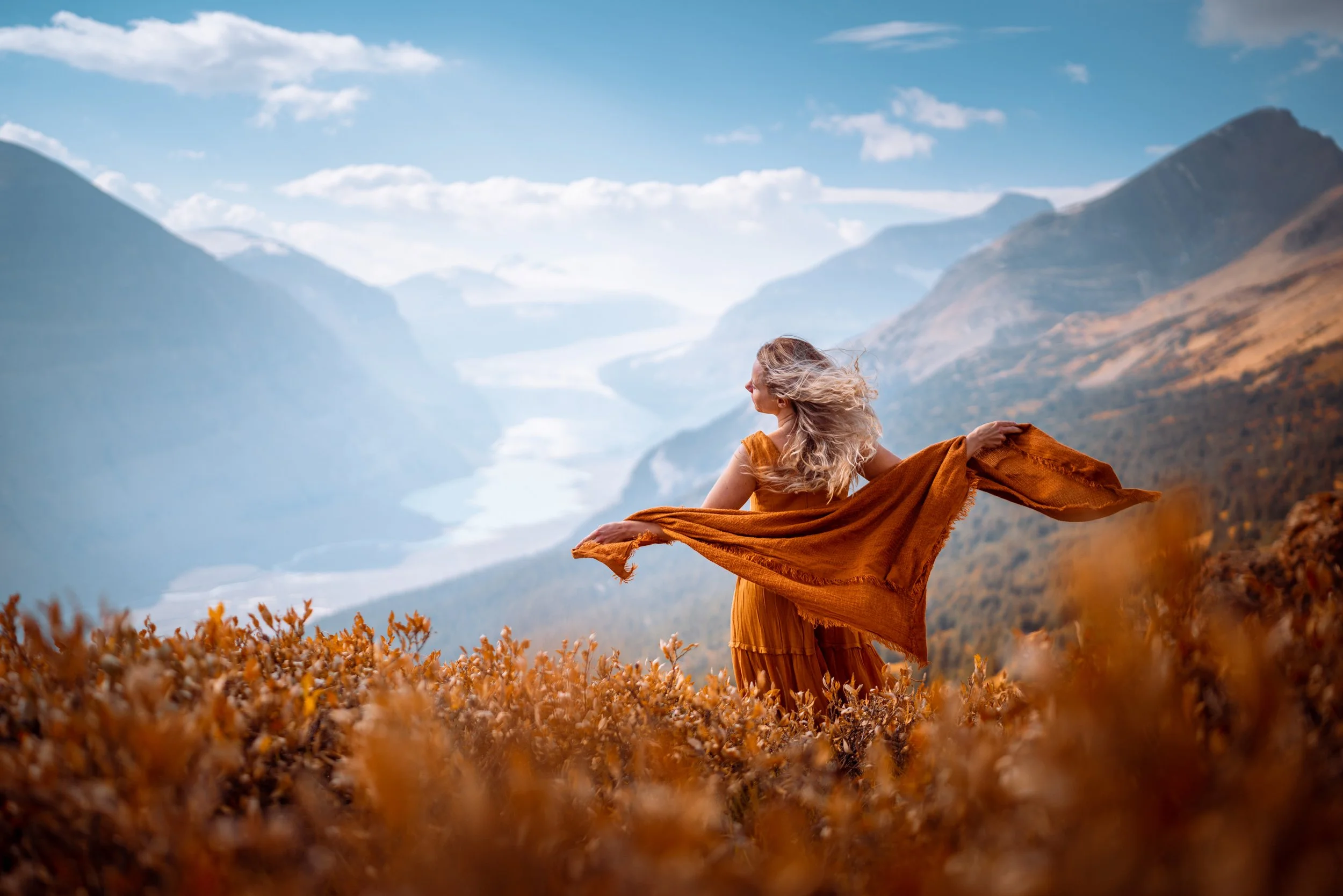 A woman in an orange dress standing in a field with mountains and blue sky in the background, holding a matching orange scarf.