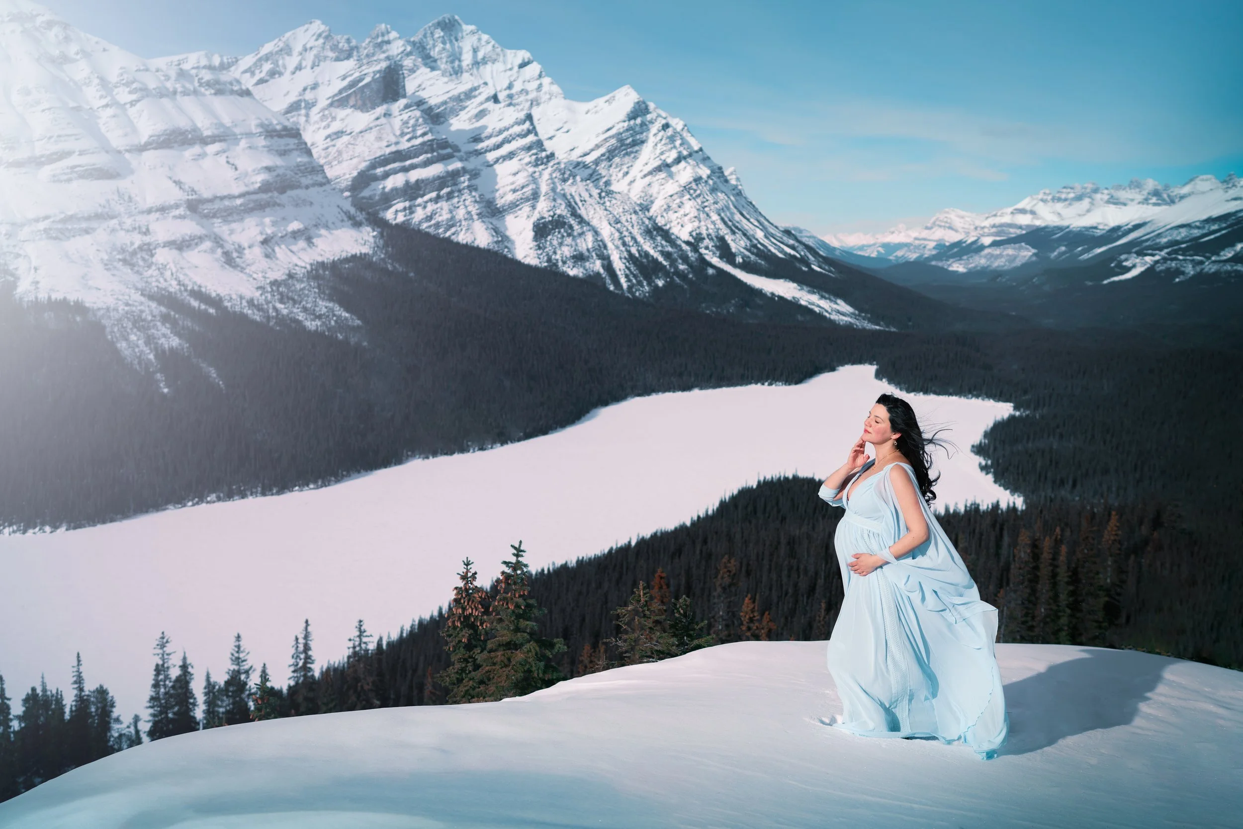 A woman in a flowing light blue dress standing on snow in a mountainous landscape with snow-covered peaks, forested slopes, and a partially frozen lake in the background.