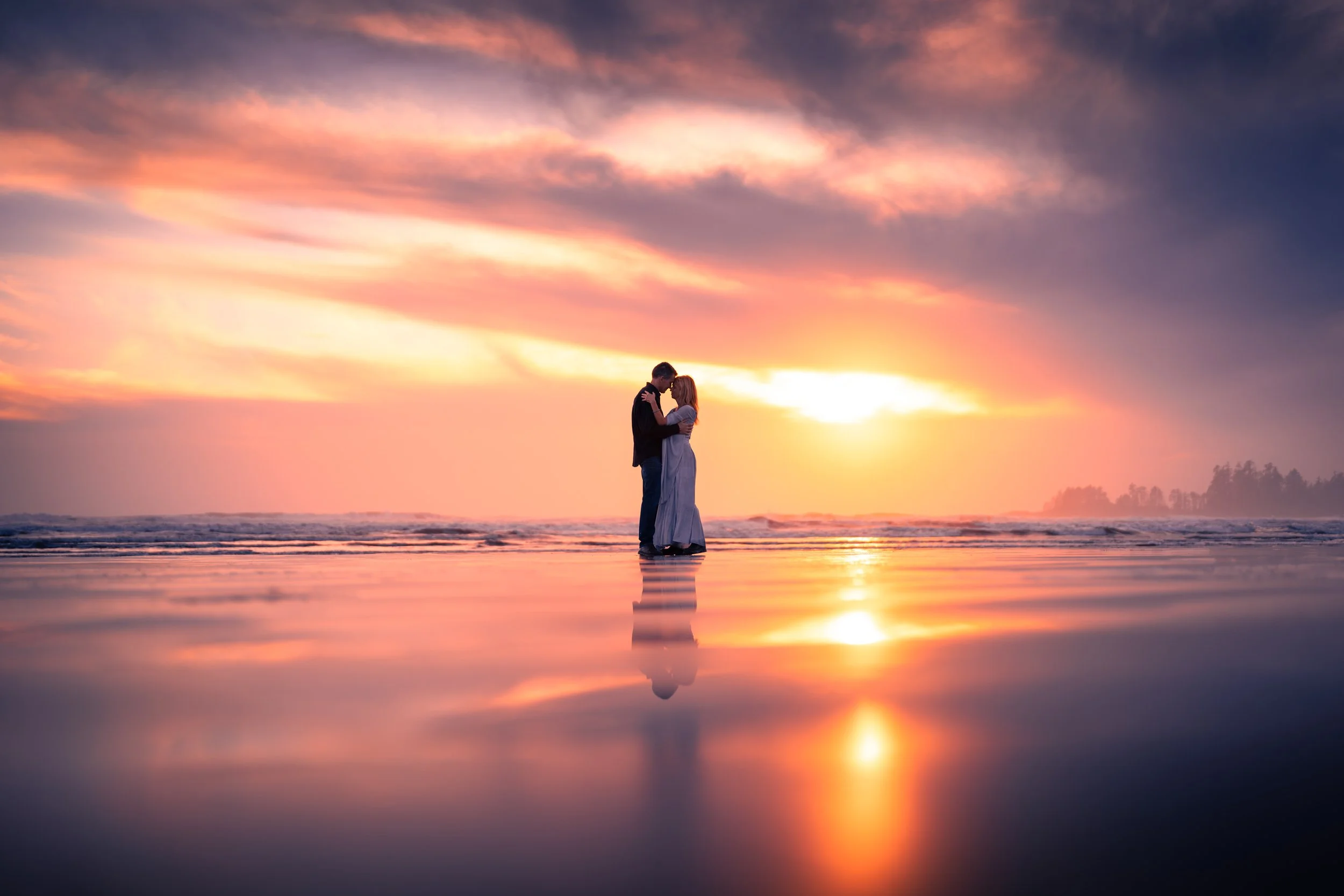 A couple standing closely on a beach at sunset, reflected on wet sand, with colorful sky and ocean waves in the background.