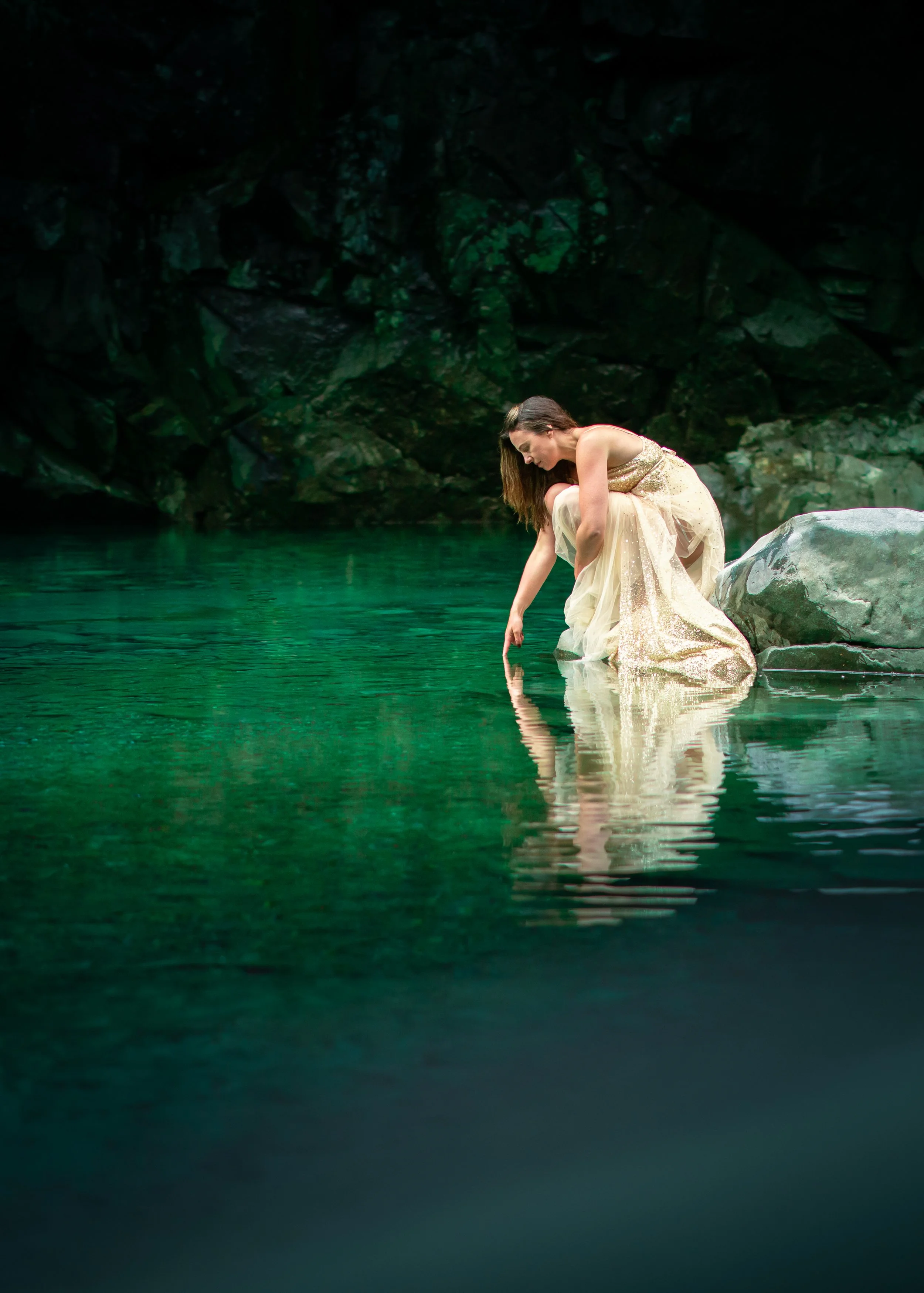 A woman in a flowing light-colored dress sits on a large rock by a calm, greenish river, touching the water with her finger. The background shows dark rocky terrain.