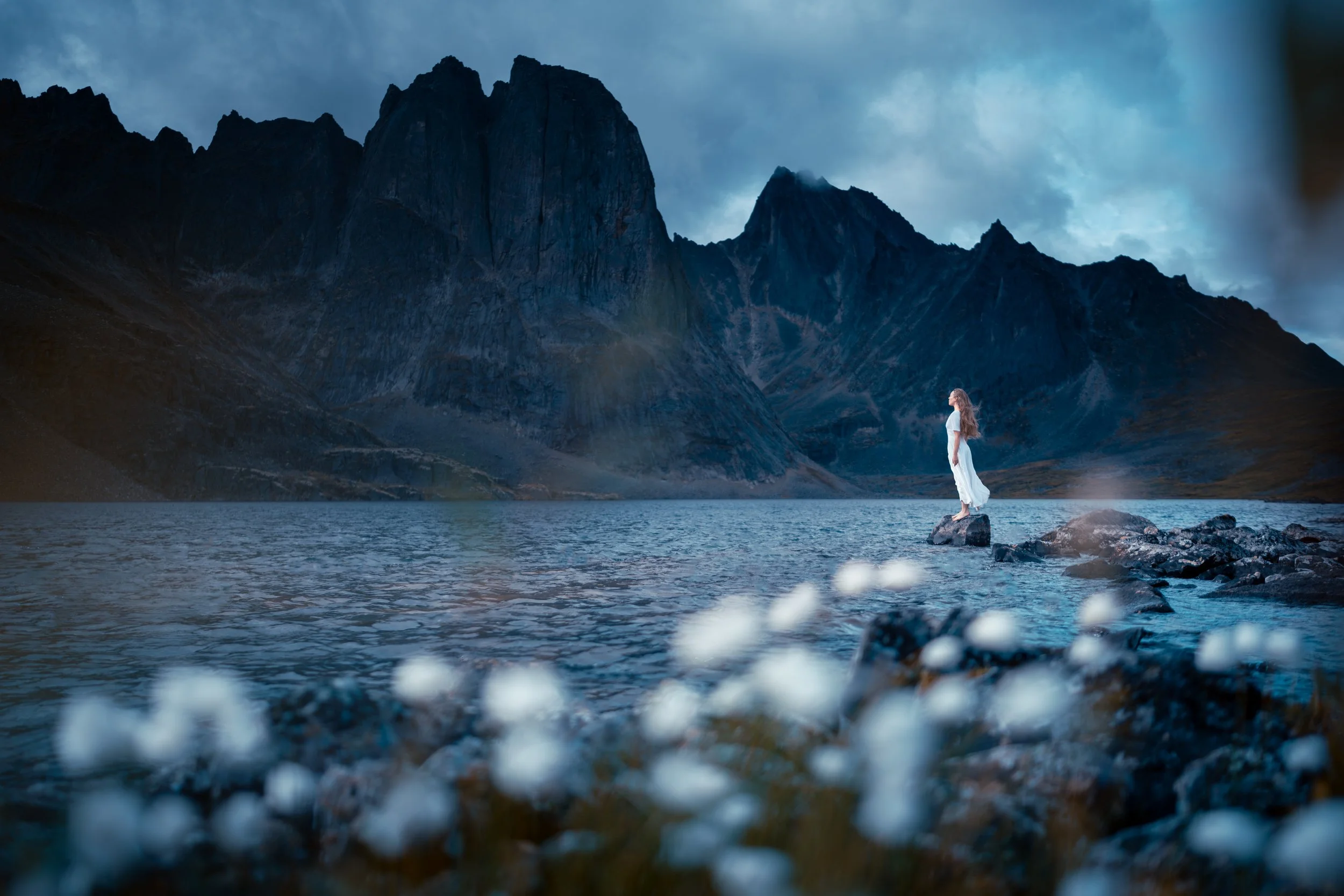 A woman in a white dress standing on a rock in a lake, with tall mountains in the background under a cloudy sky.
