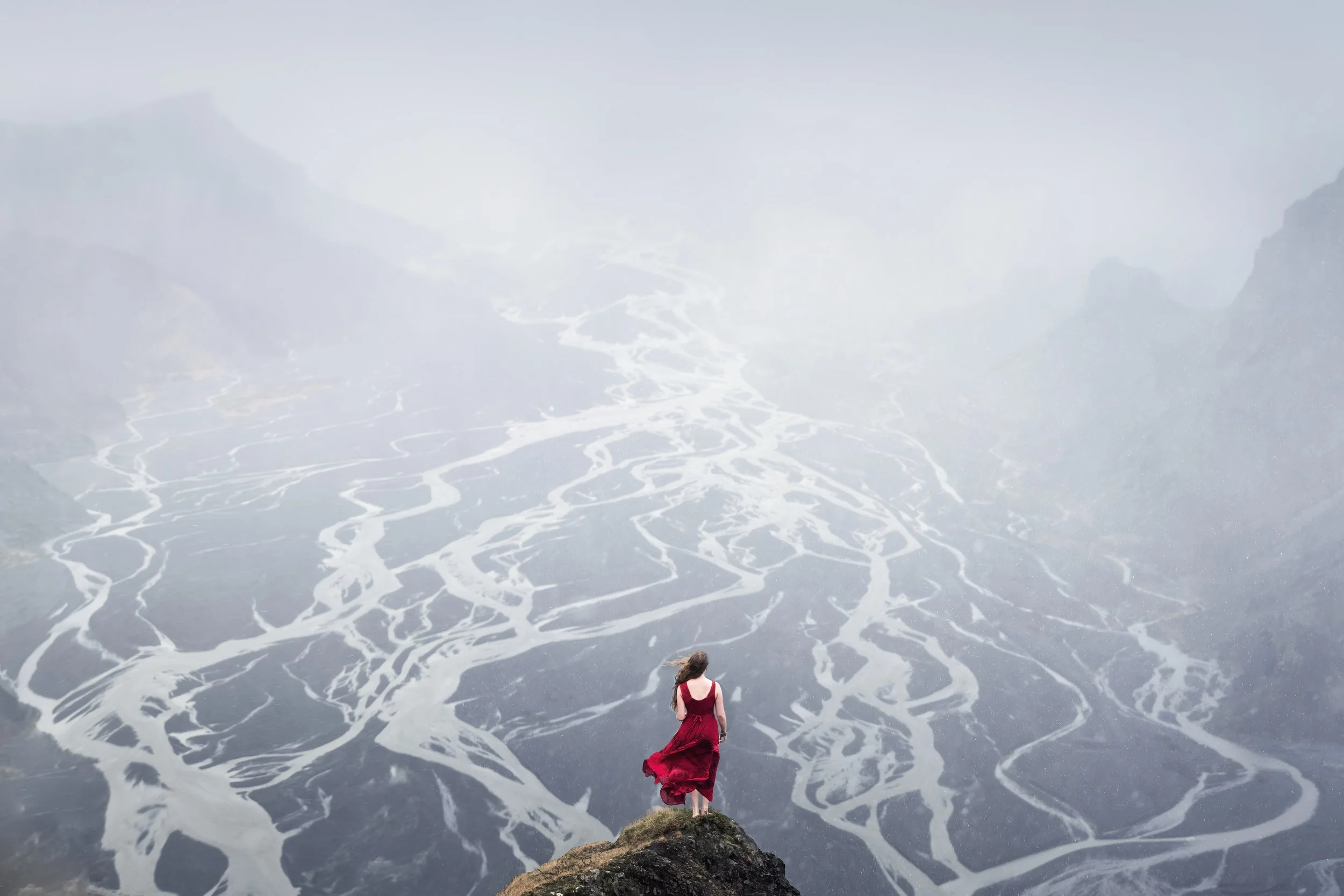 A woman in a red dress stands on the edge of a cliff, overlooking a river valley with winding streams and mountains shrouded in fog in the background.