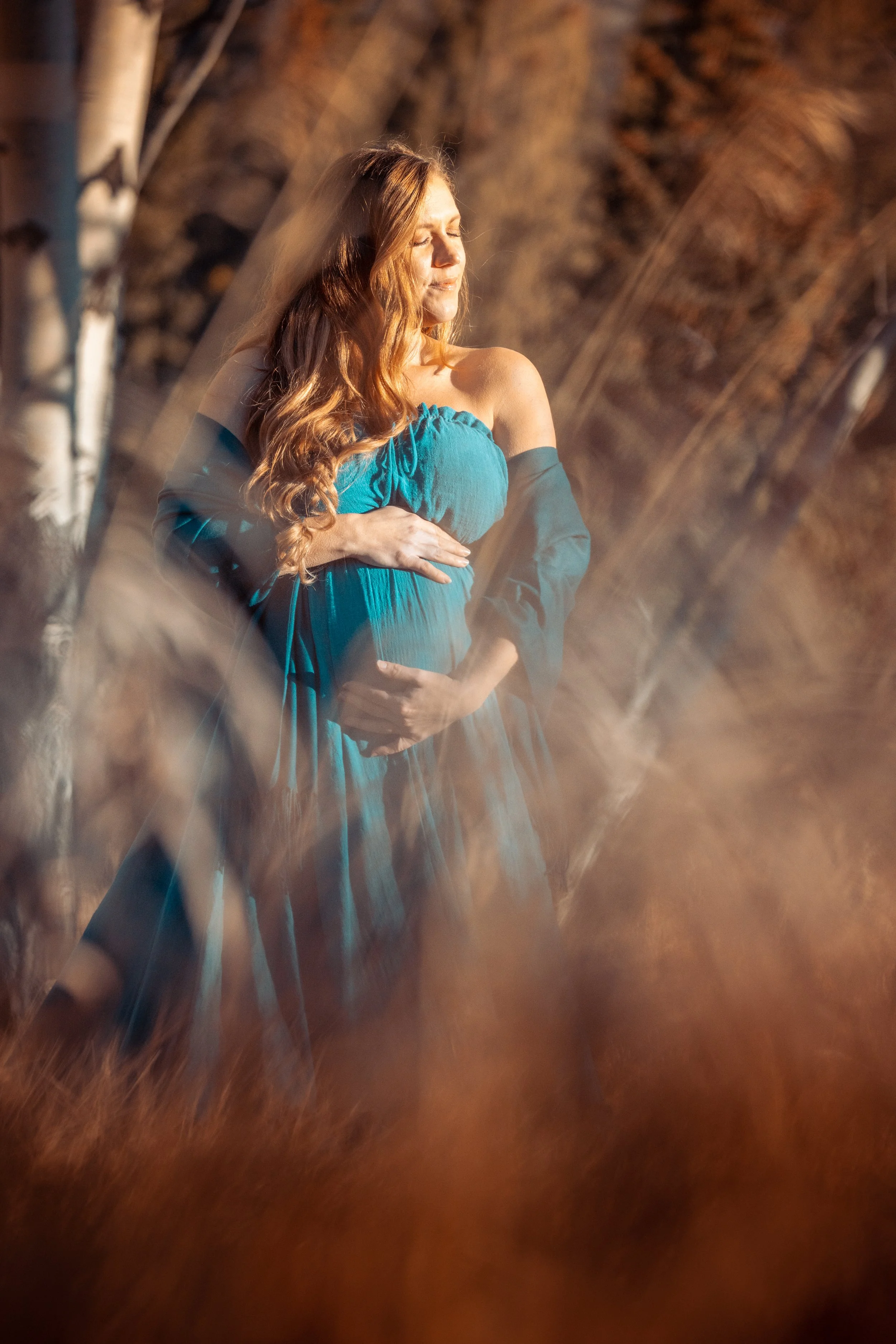 A pregnant woman with long, wavy red hair, wearing a blue off-the-shoulder dress, stands outdoors with her eyes closed, gently touching her belly as sunlight illuminates her face.
