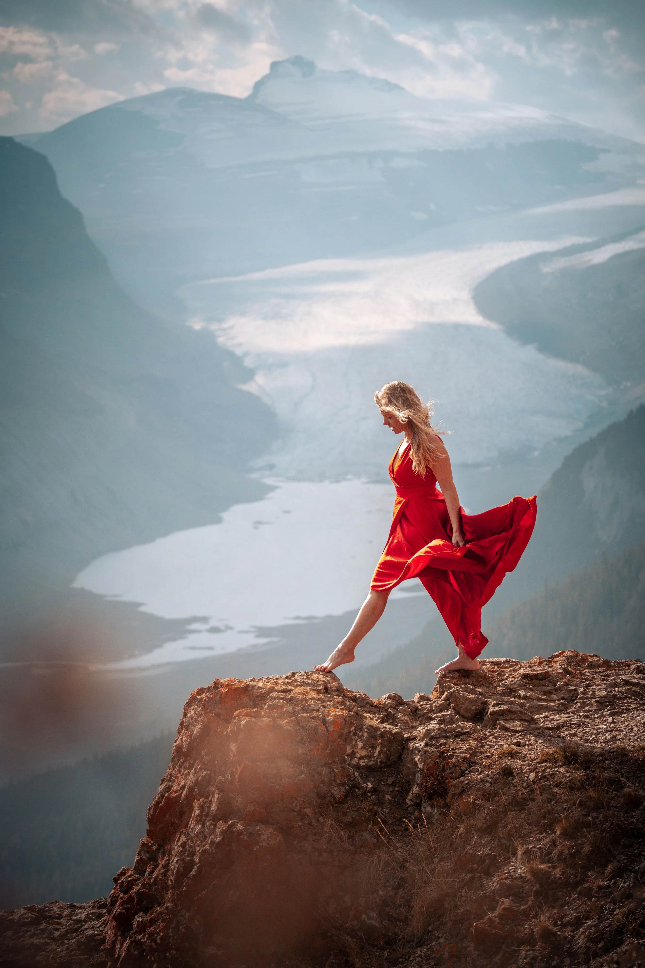 A woman in a red dress standing on a rocky cliff overlooking a vast glacier with mountains and ice in the background.