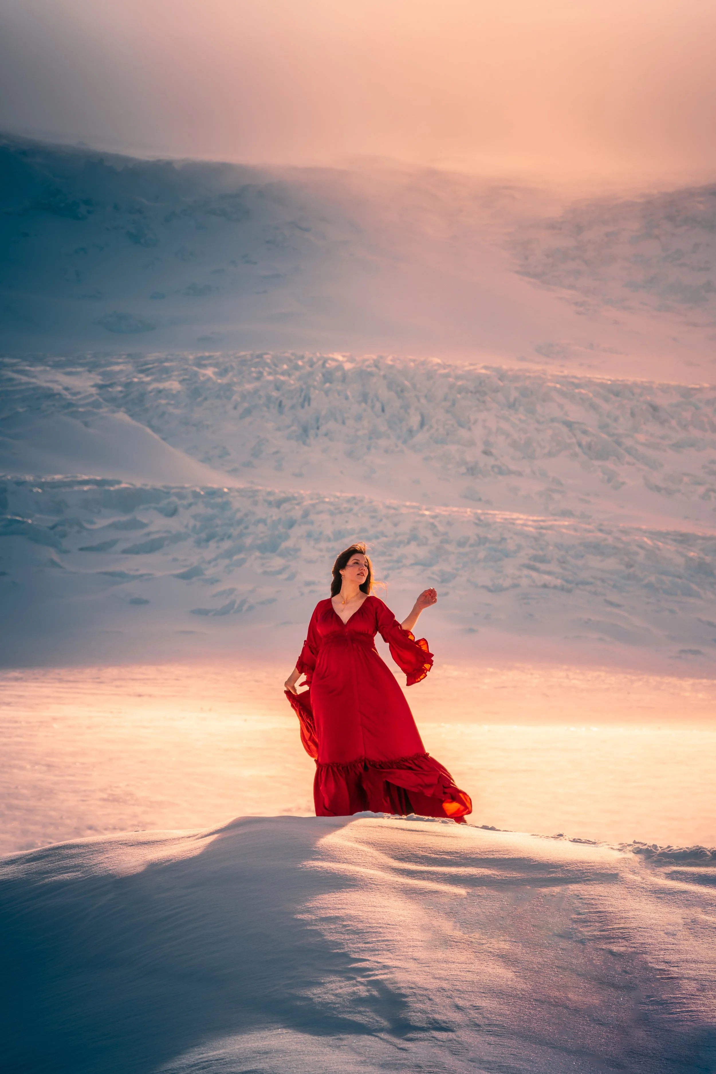 A woman in a long red dress standing on snow-covered ground with a backdrop of a sunset sky and snow-covered mountains.