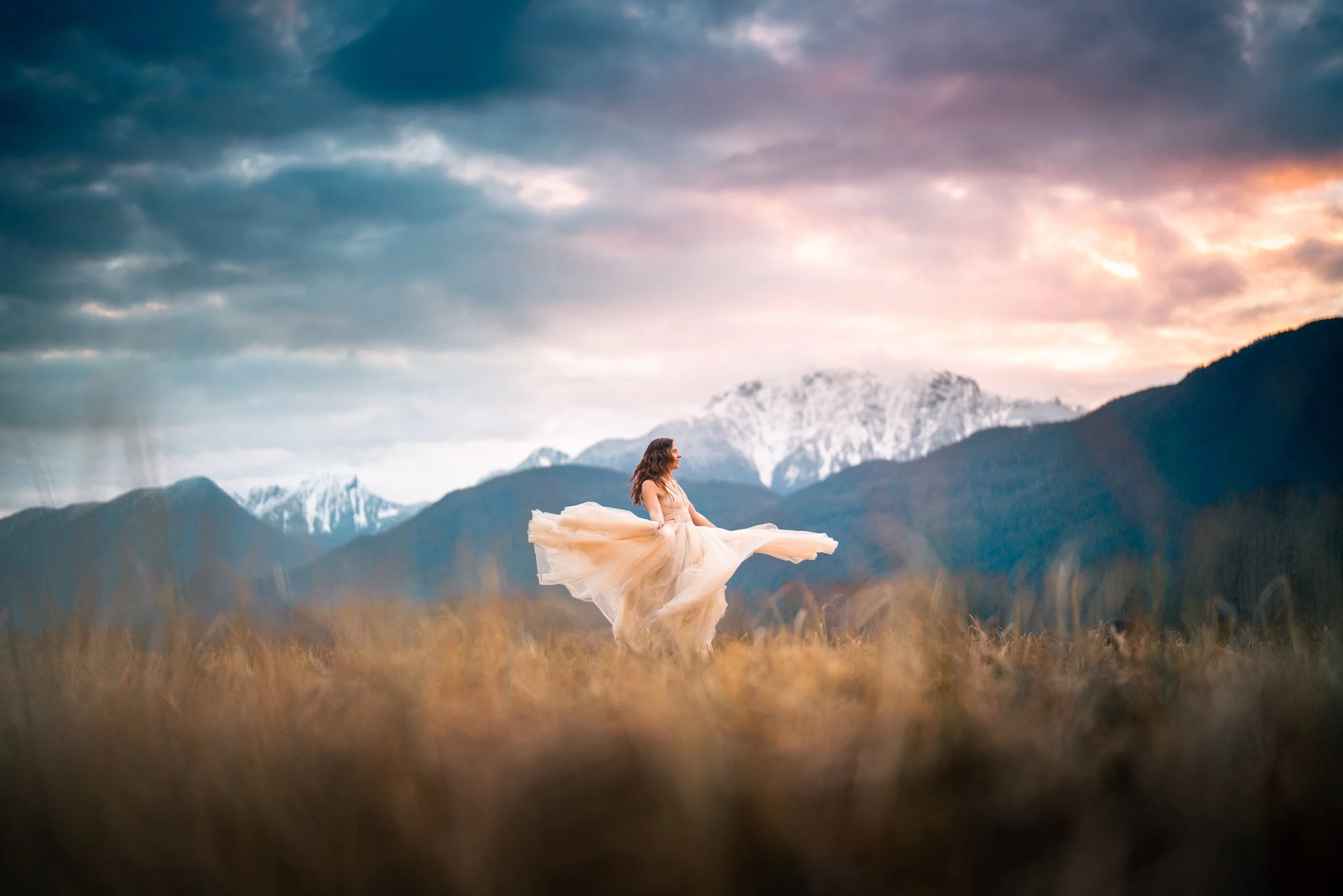 A woman in a flowing dress standing in a field with mountains and a dramatic sky in the background.