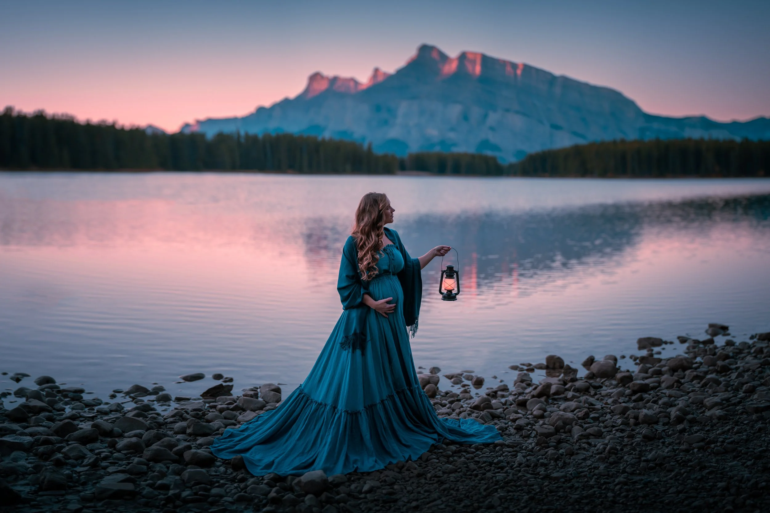 Pregnant woman in a flowing blue gown standing on rocky shore, holding a lantern, by a calm lake with mountain backdrop at sunset