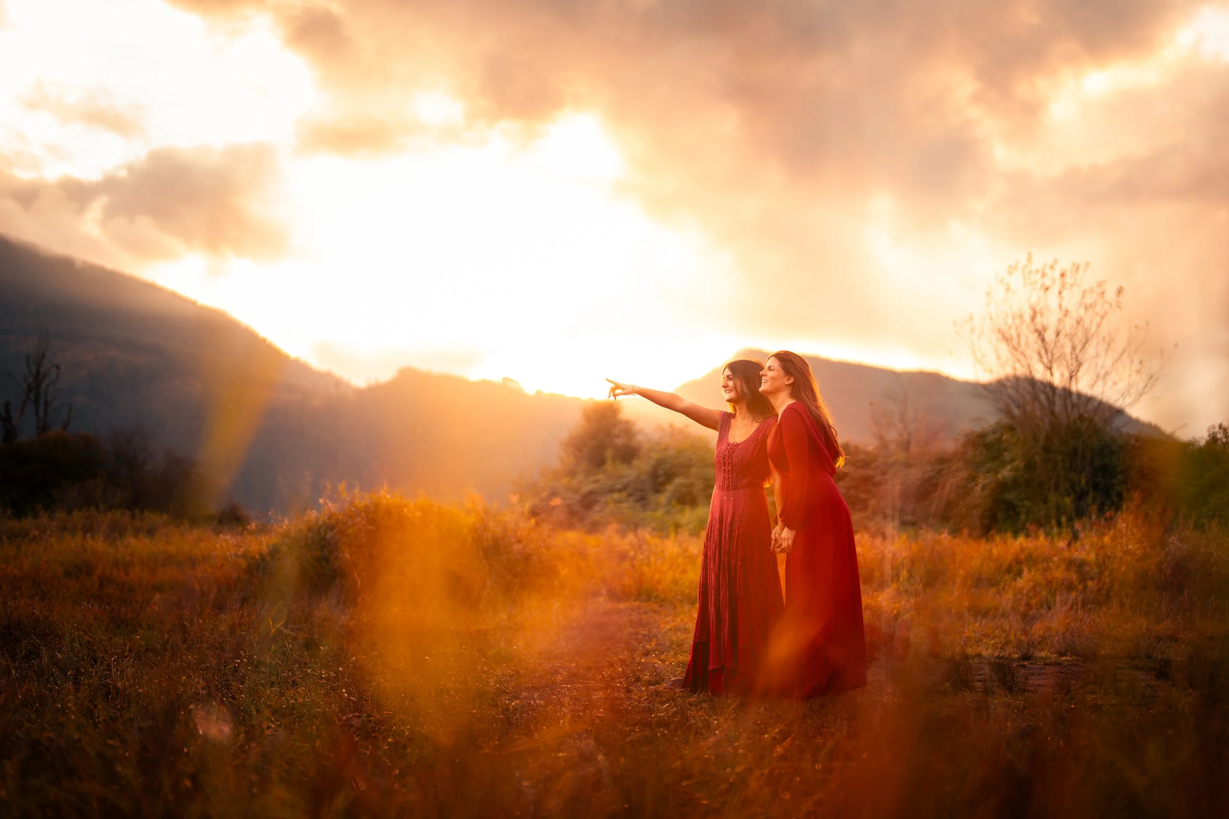 Two women in red dresses standing hand in hand in a field at sunset, one pointing towards the sky with a mountain in the background and a dramatic, fiery sky.