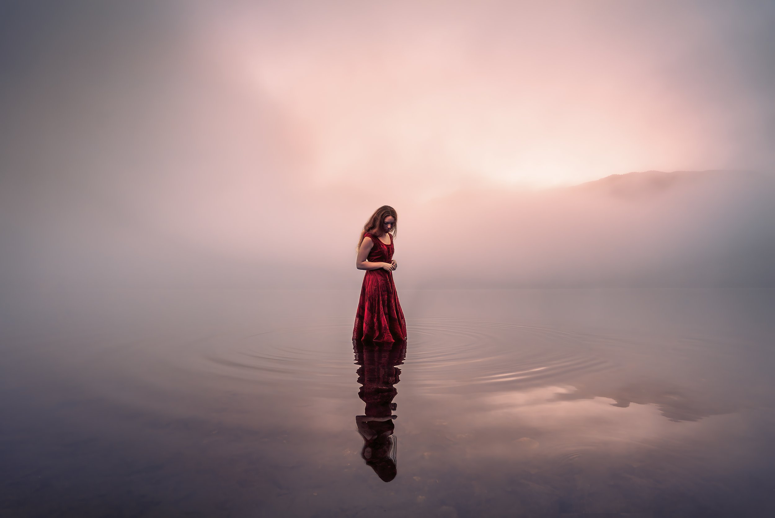 A woman in a red dress standing in calm water, with a serene, foggy pink sky behind her.