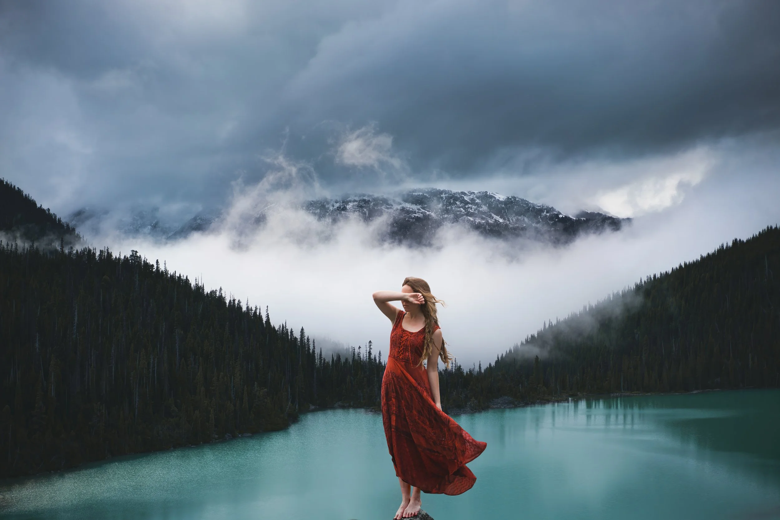 A woman in a red dress standing on a rock by a teal-colored lake, with foggy mountains and dense pine forests in the background under a cloudy sky.