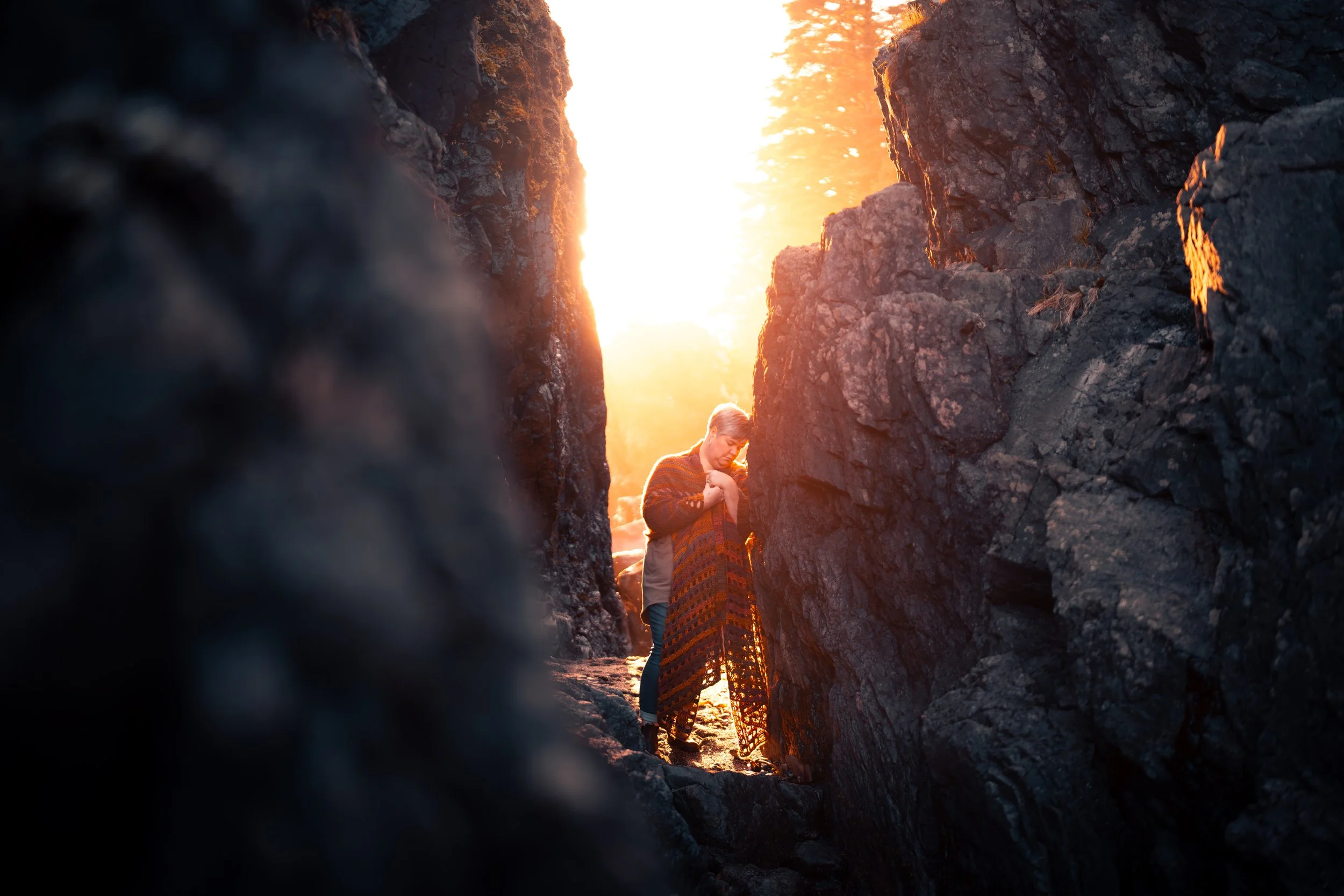 Person standing between tall, dark rock formations at sunset, holding a colorful blanket.