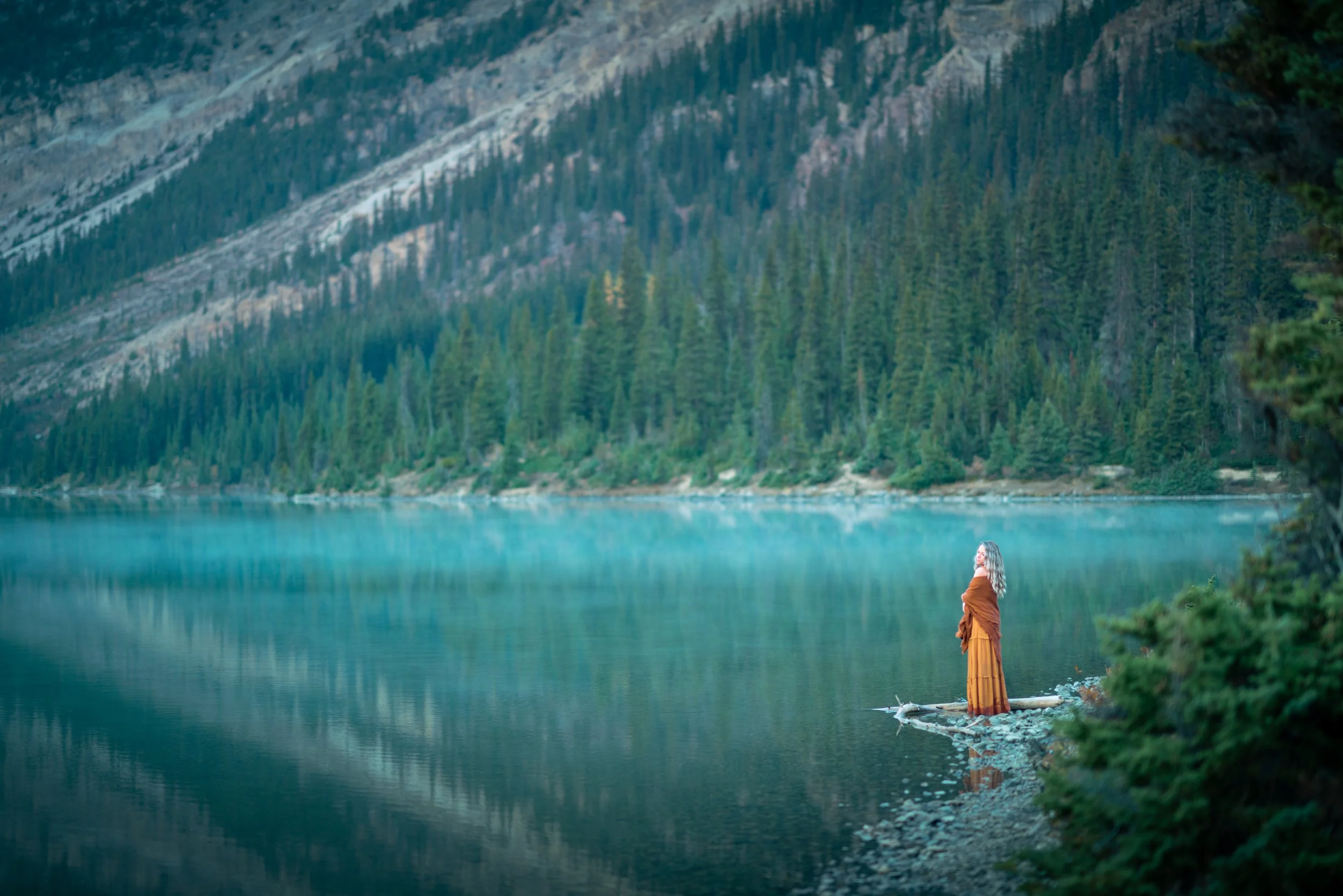 A woman stands on a small rocky shore beside a calm mountain lake, surrounded by dense evergreen forest and steep mountain slopes in the background.