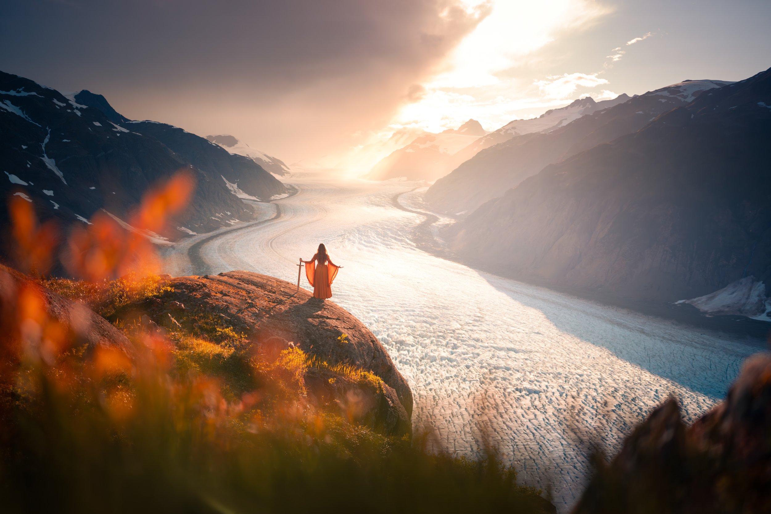 A person standing on a rocky hilltop overlooking a glacier in a mountain valley at sunset.