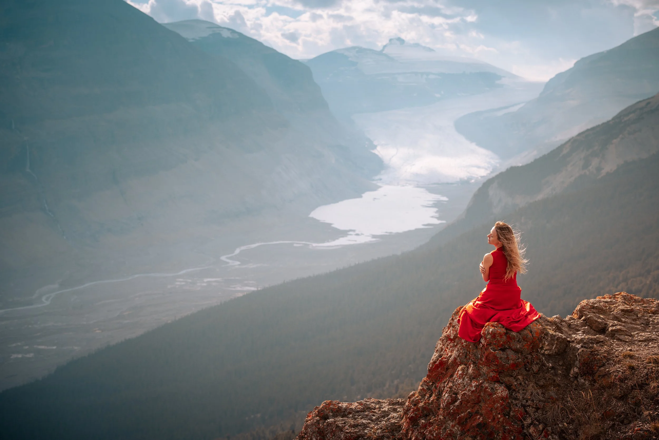 A woman in a red dress sitting on a rocky ledge overlooking a vast valley with a winding river and mountains in the background.
