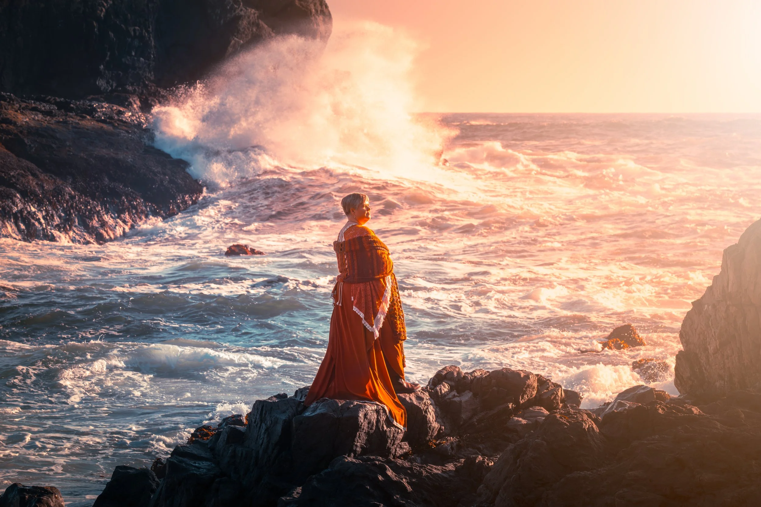 A woman in historical dress standing on rocks by the ocean at sunset, waves crashing against rocks, warm orange glow in the sky.