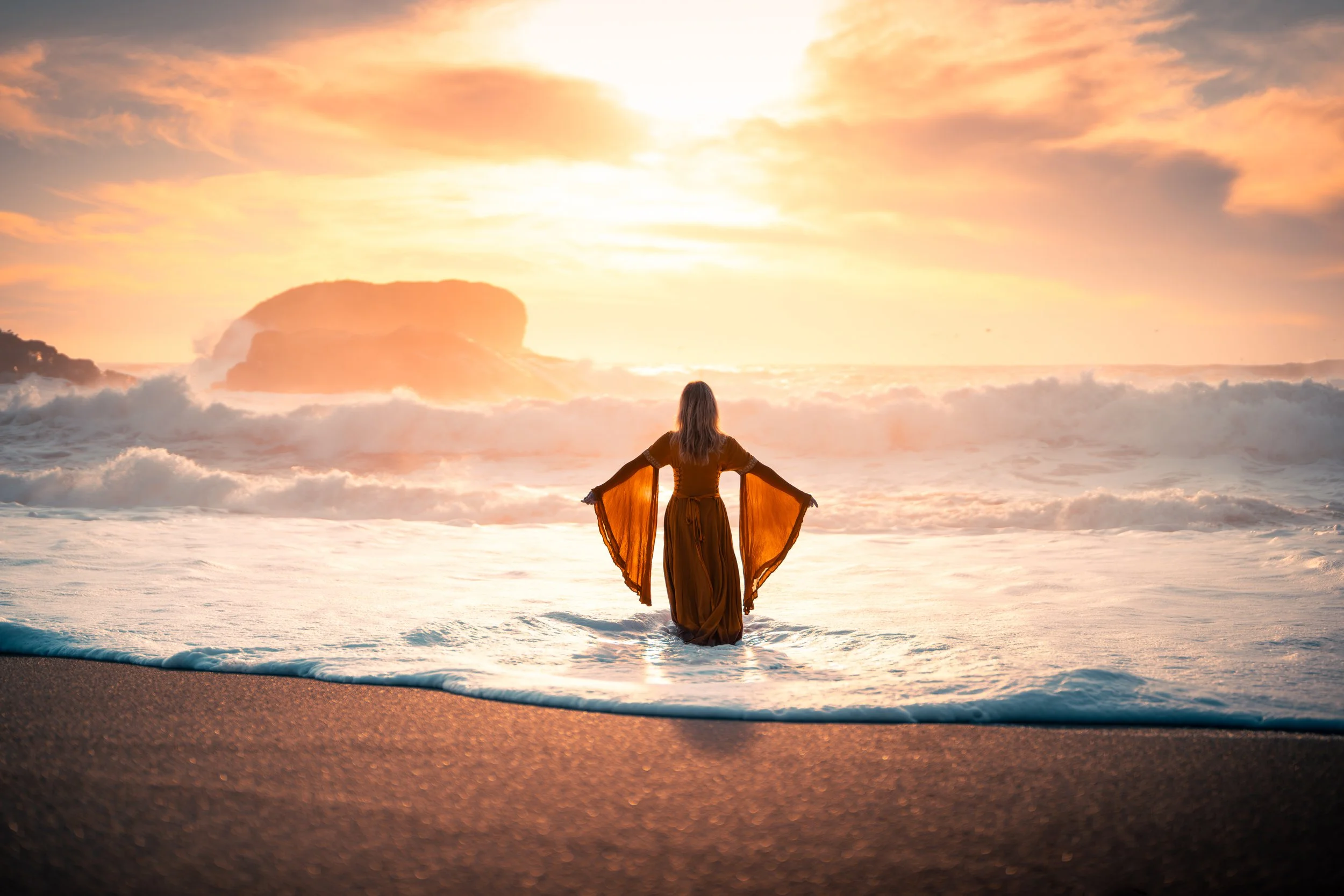 A woman standing in the ocean waves during sunset, with her arms outstretched and wearing a flowing dress.