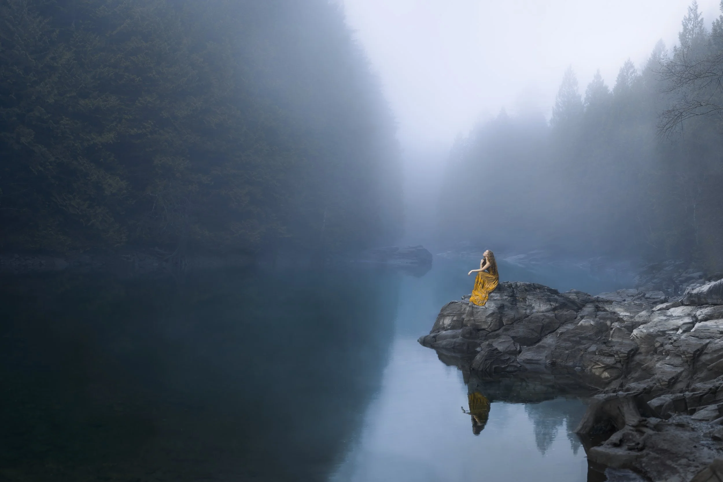 A woman in a yellow dress sitting on a large rock by a calm, reflective river surrounded by fog and dense trees.