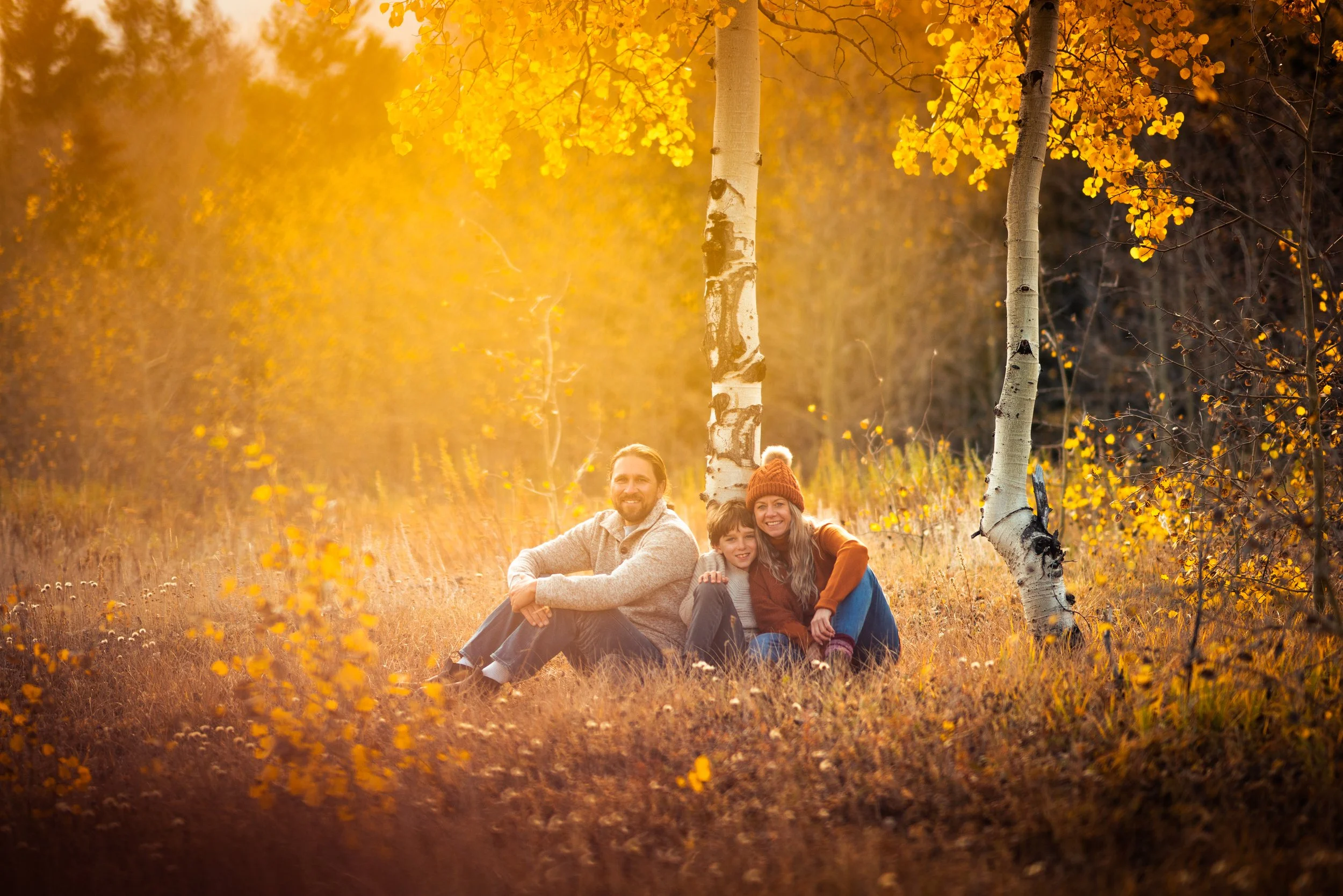 A family of three sitting on the ground in a forest with autumn leaves and trees, smiling and enjoying the sunshine.