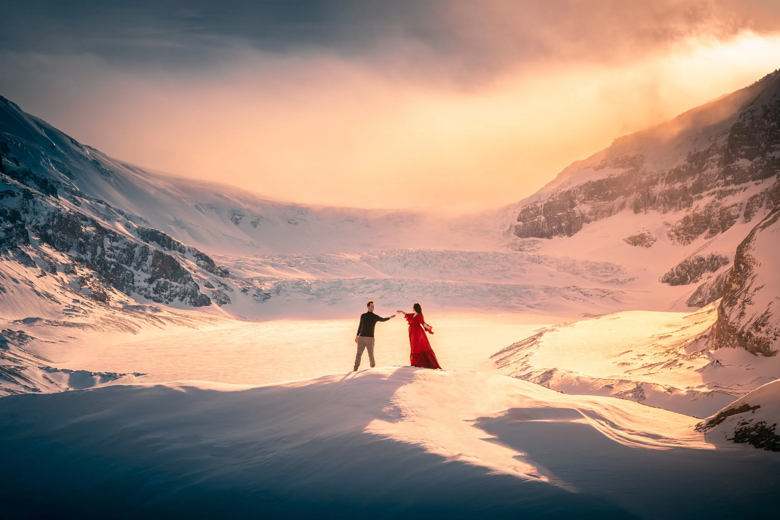 A couple holds hands in a snowy mountain landscape during sunset, with snow-covered peaks and a glowing sky.