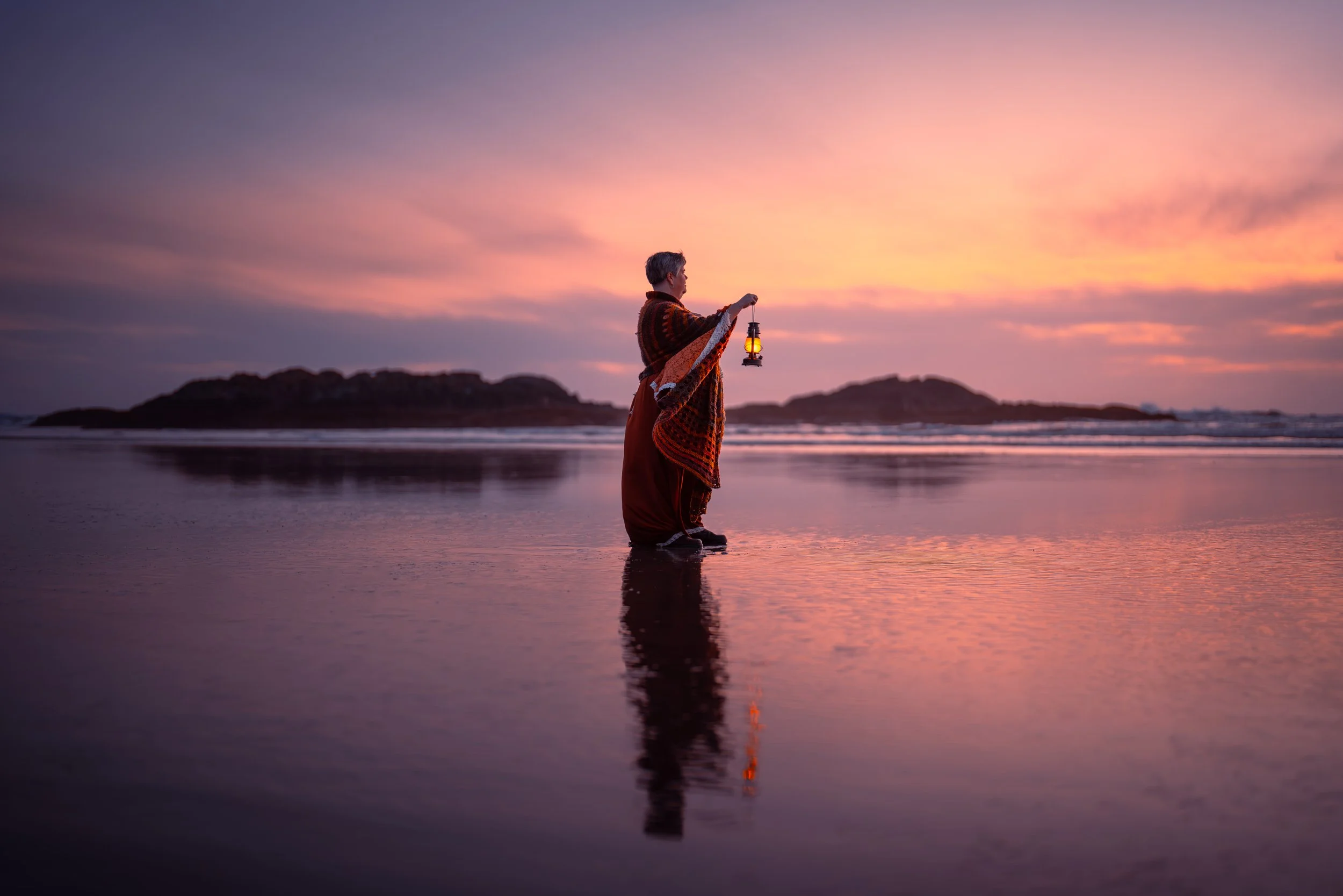 Person standing in shallow water at sunset, holding a lantern, with pink and purple sky and distant islands.