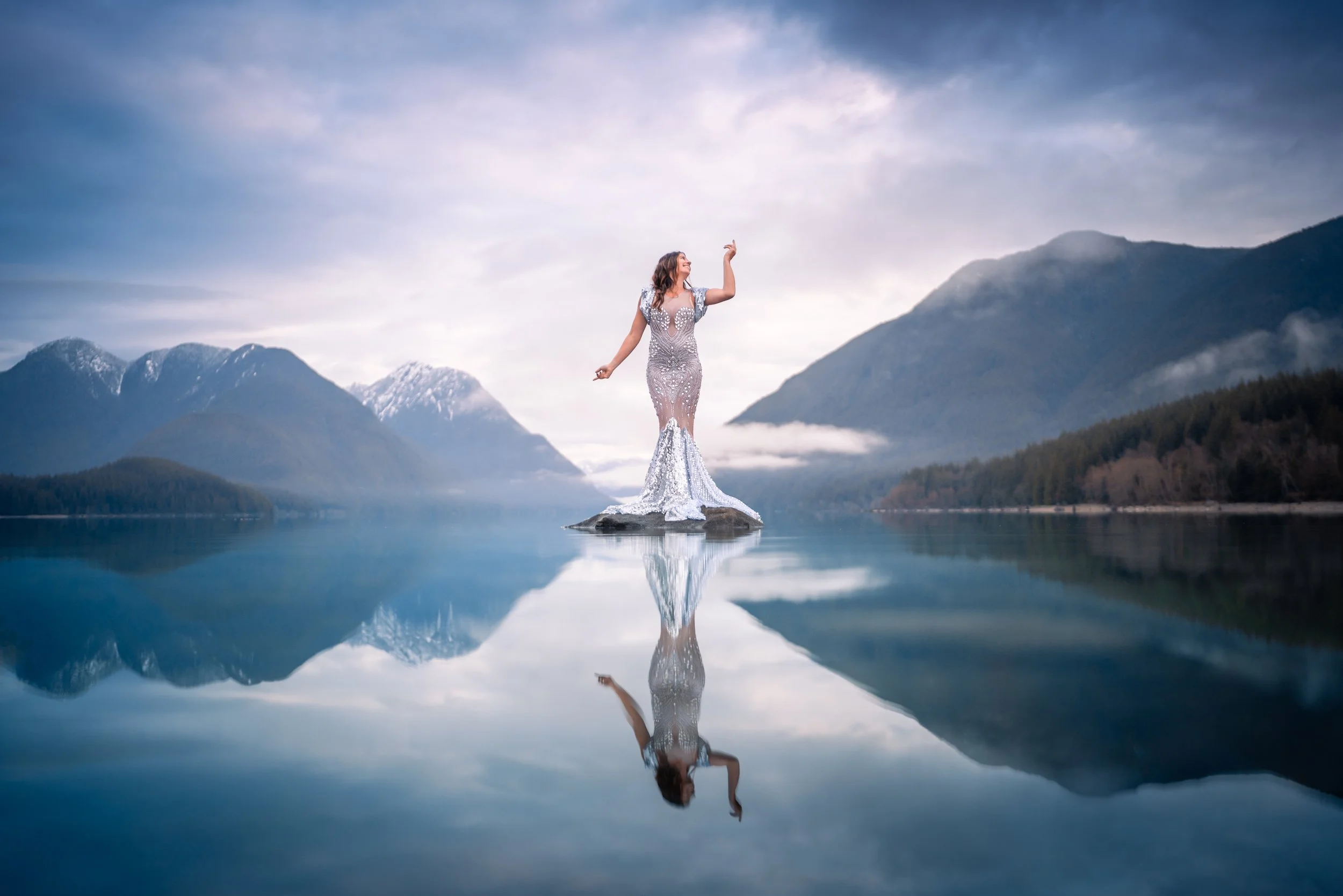 A woman in a silver evening gown standing on a rock in a calm lake, with mountains and cloudy sky in the background, and her reflection visible on the water.