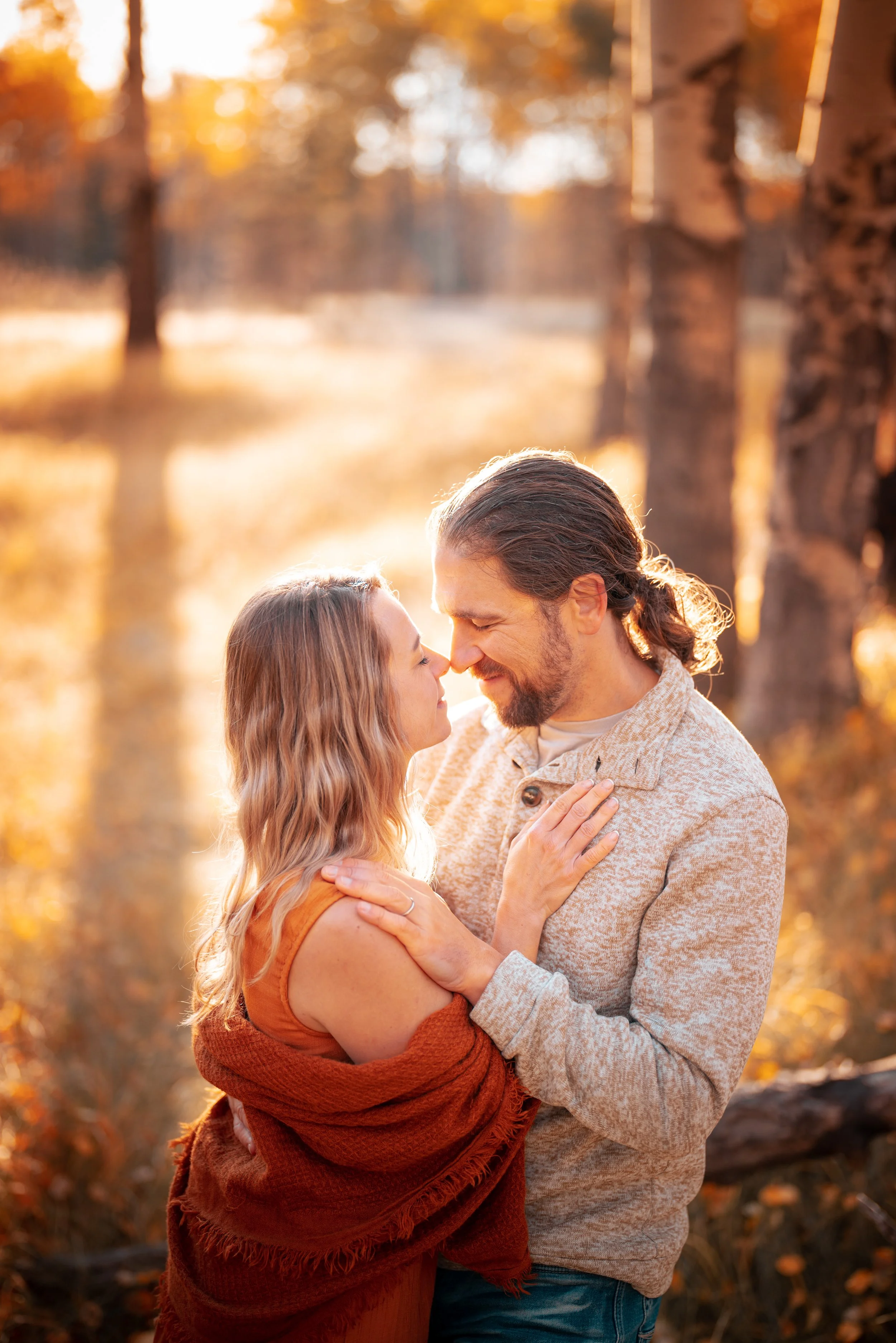 A couple embracing outdoors during autumn sunset, with trees and water in background.