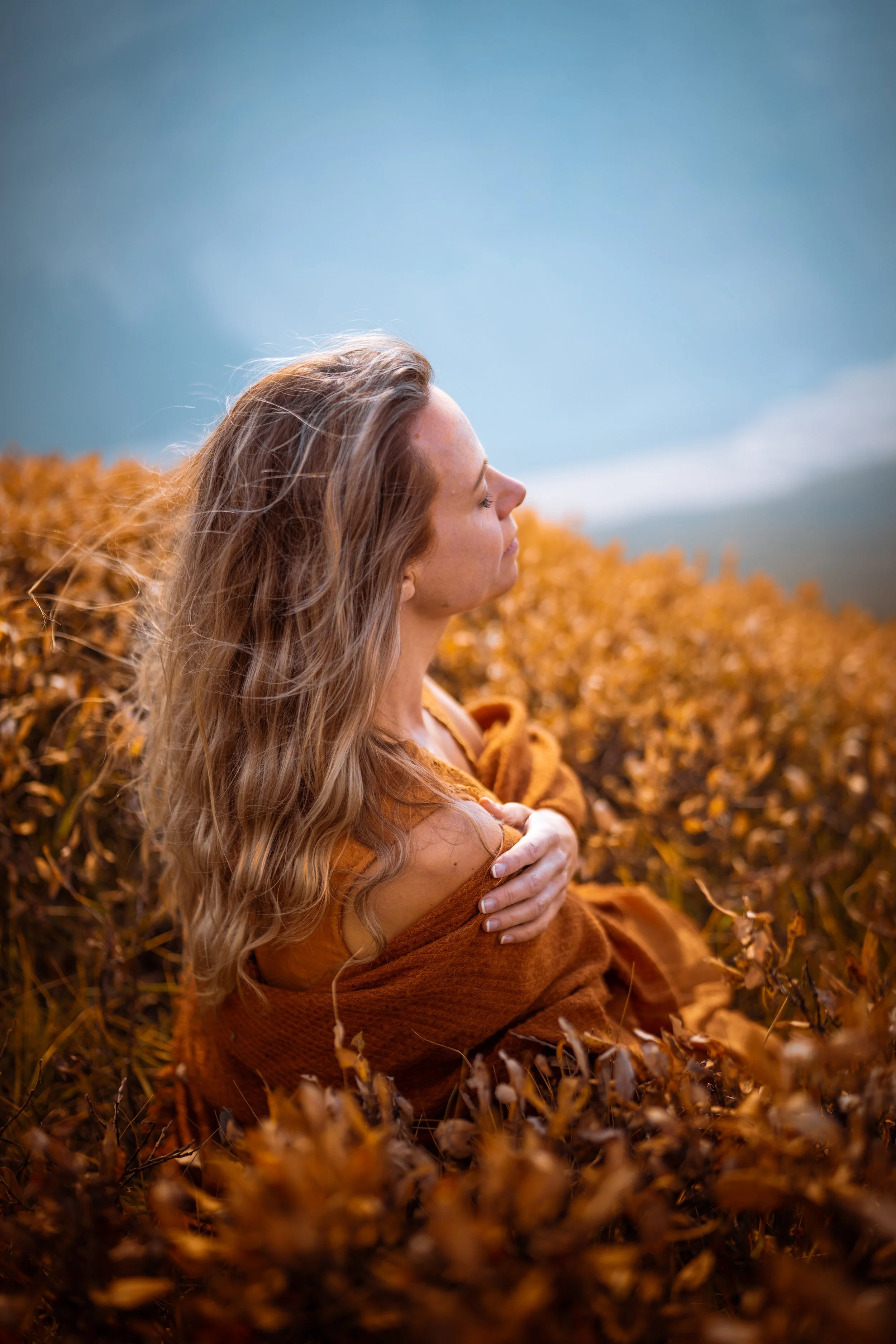 A woman with long wavy hair, sitting among autumn-colored bushes with her eyes closed and arms crossed over her chest, embracing herself on a cloudy day.