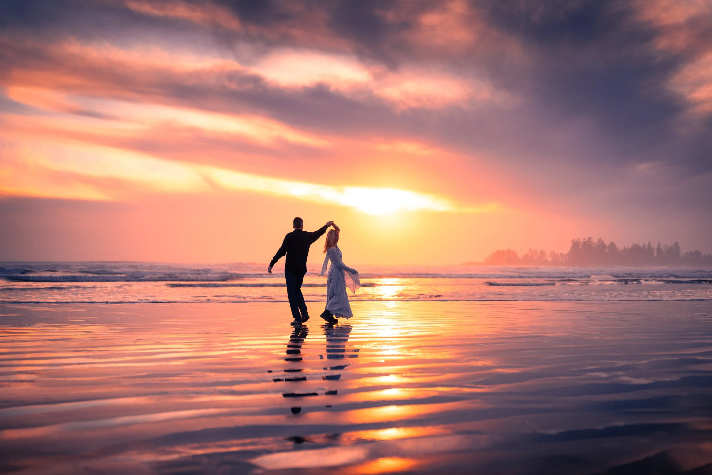 A couple dancing on the beach during sunset, with a colorful sky and calm ocean in the background.