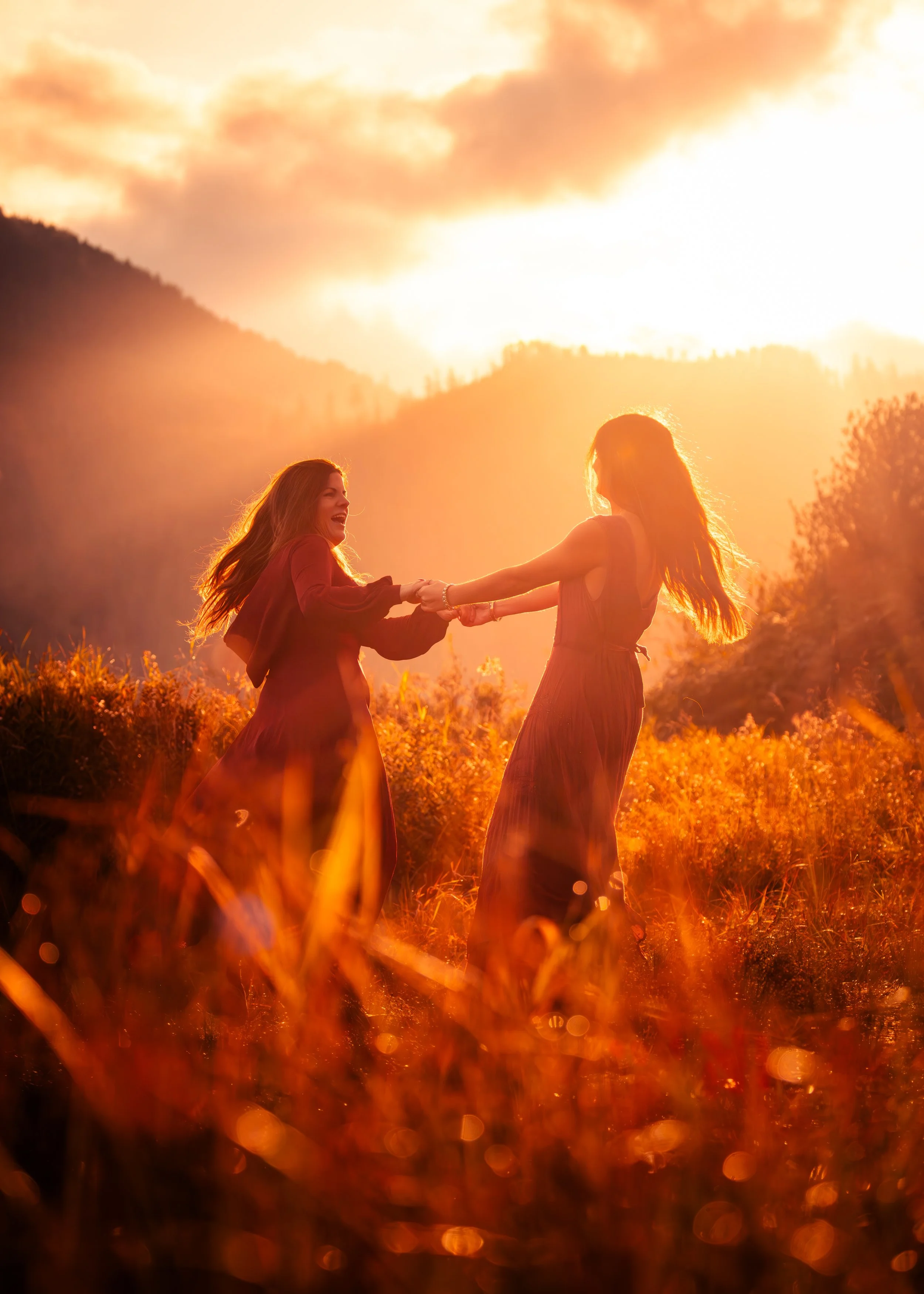 Two women are holding hands and spinning in a field during sunset, with the sky glowing orange and mountains in the background.
