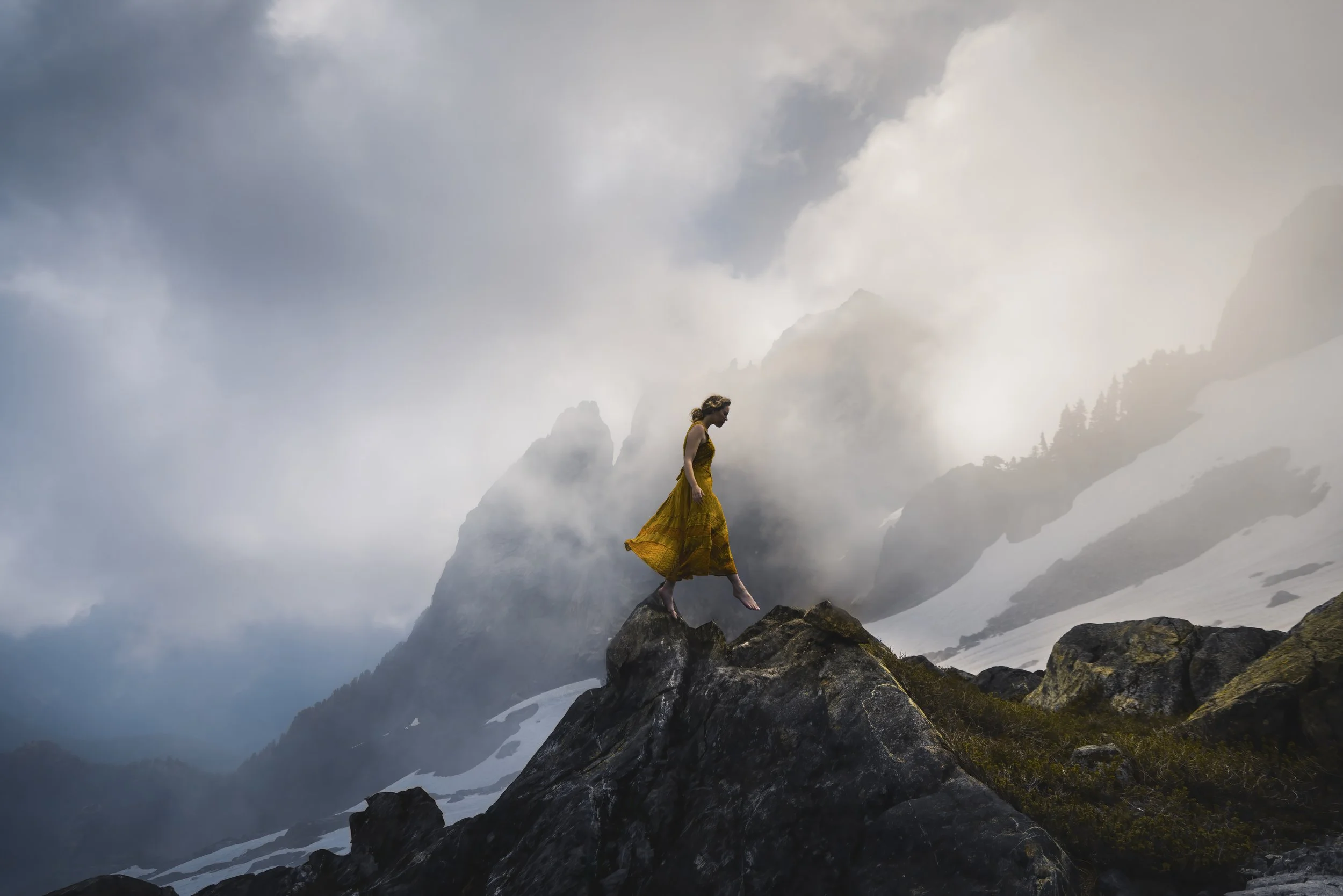 A woman in a yellow dress walking on a large rock peak with a mountainous, cloudy, and snowy landscape in the background.