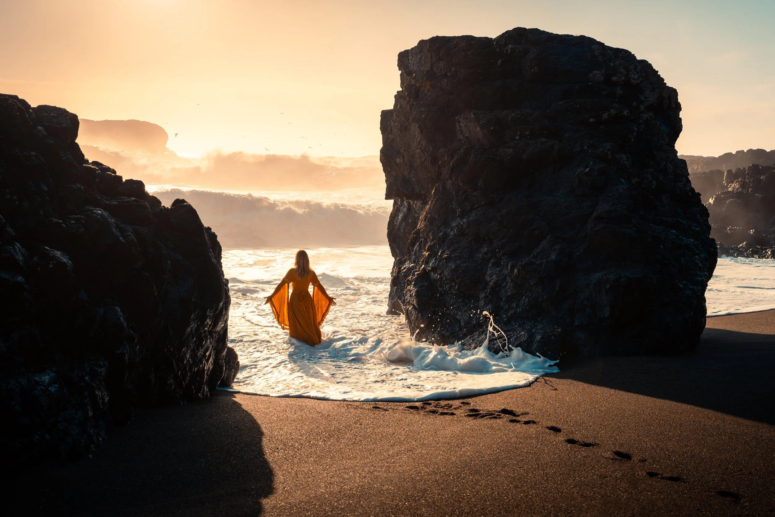 A woman in a long, flowing yellow dress stands at the edge of the ocean between two large black rocks during a sunset or sunrise.