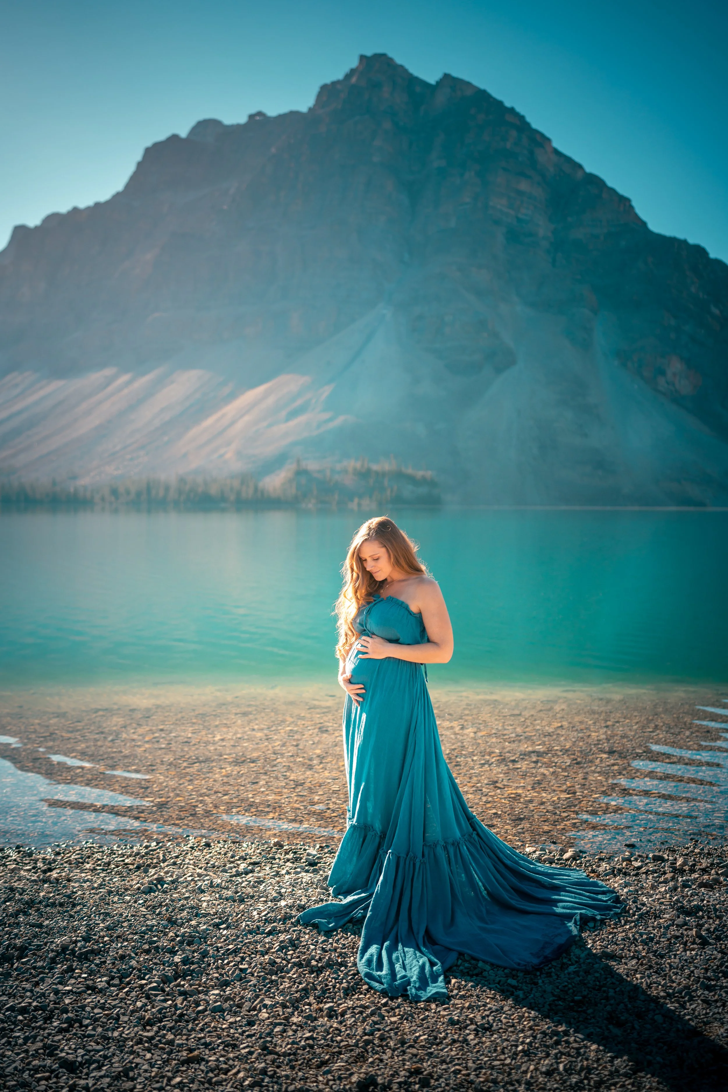 Pregnant woman in a flowing blue dress standing on rocky shoreline with a mountain and lake in the background.