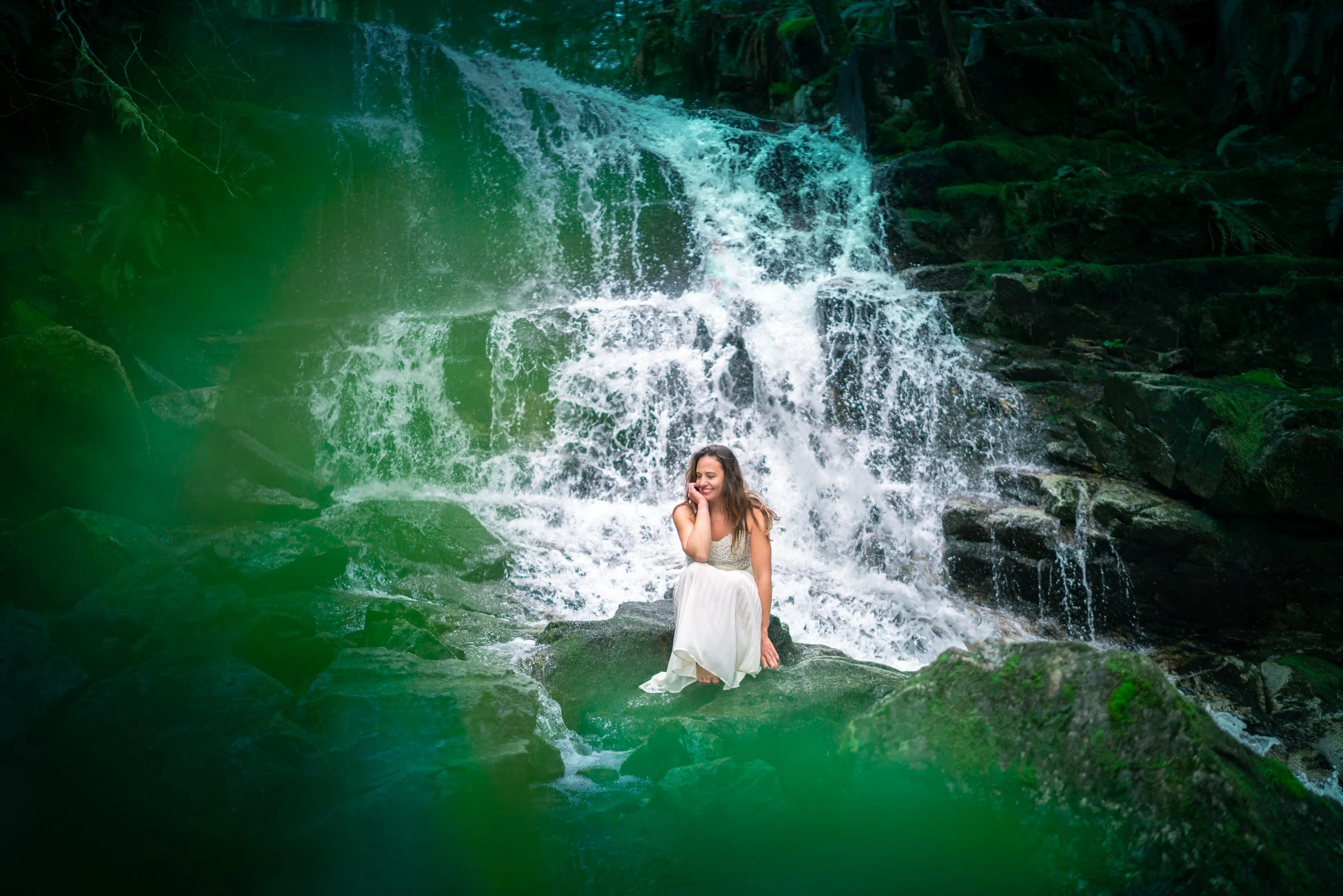 A woman in a white dress sitting on a rock in front of a waterfall surrounded by lush green foliage.